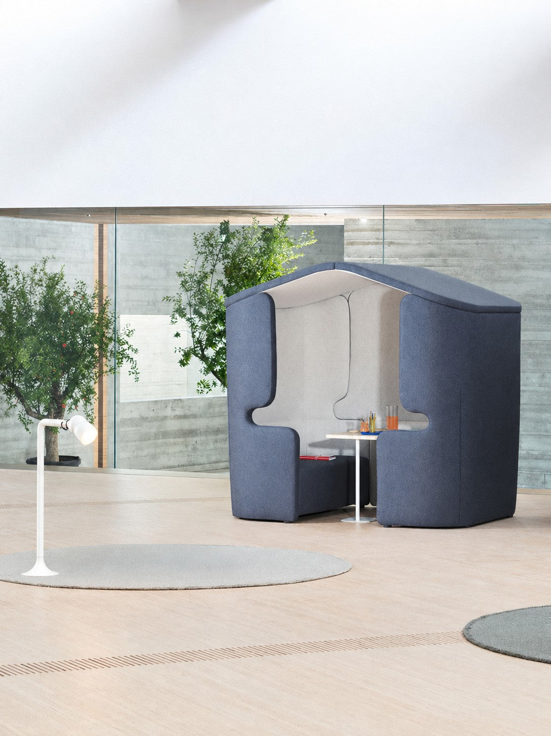 Un espace de travail ouvert et moderne avec l'Alcôve acoustique Zen 4 pers bicolore + table blanche de Monsieur Bureau, posé sur un sol en bois clair avec des plantes devant de grandes baies vitrées et un lampadaire blanc à proximité.