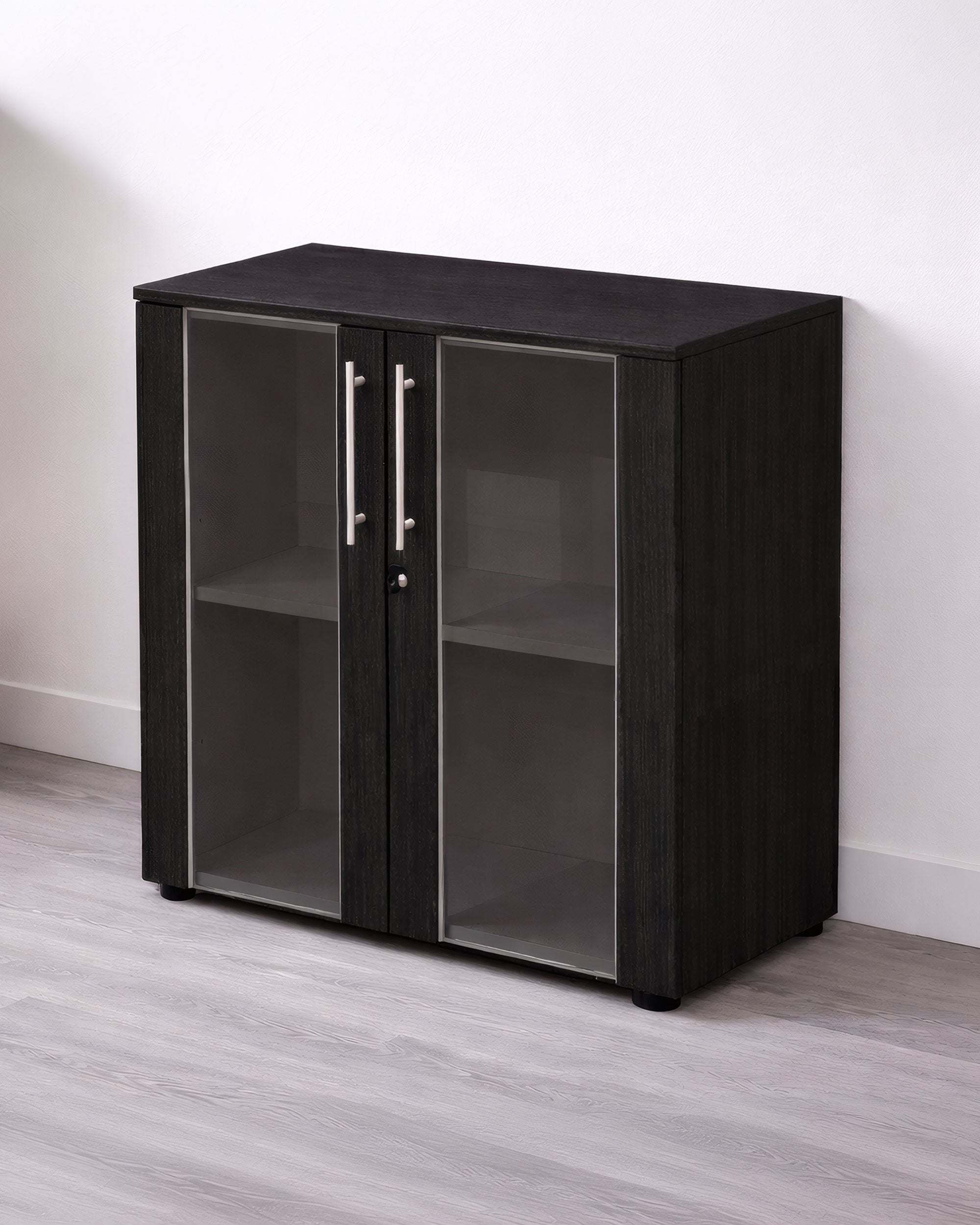 L'armoire Monsieur Bureau Basse Flora Portes Vitrées H81.4 cm comporte deux portes en verre avec des poignées argentées et deux étagères pour un rangement professionnel, présentée contre un mur blanc sur un sol en bois gris clair.