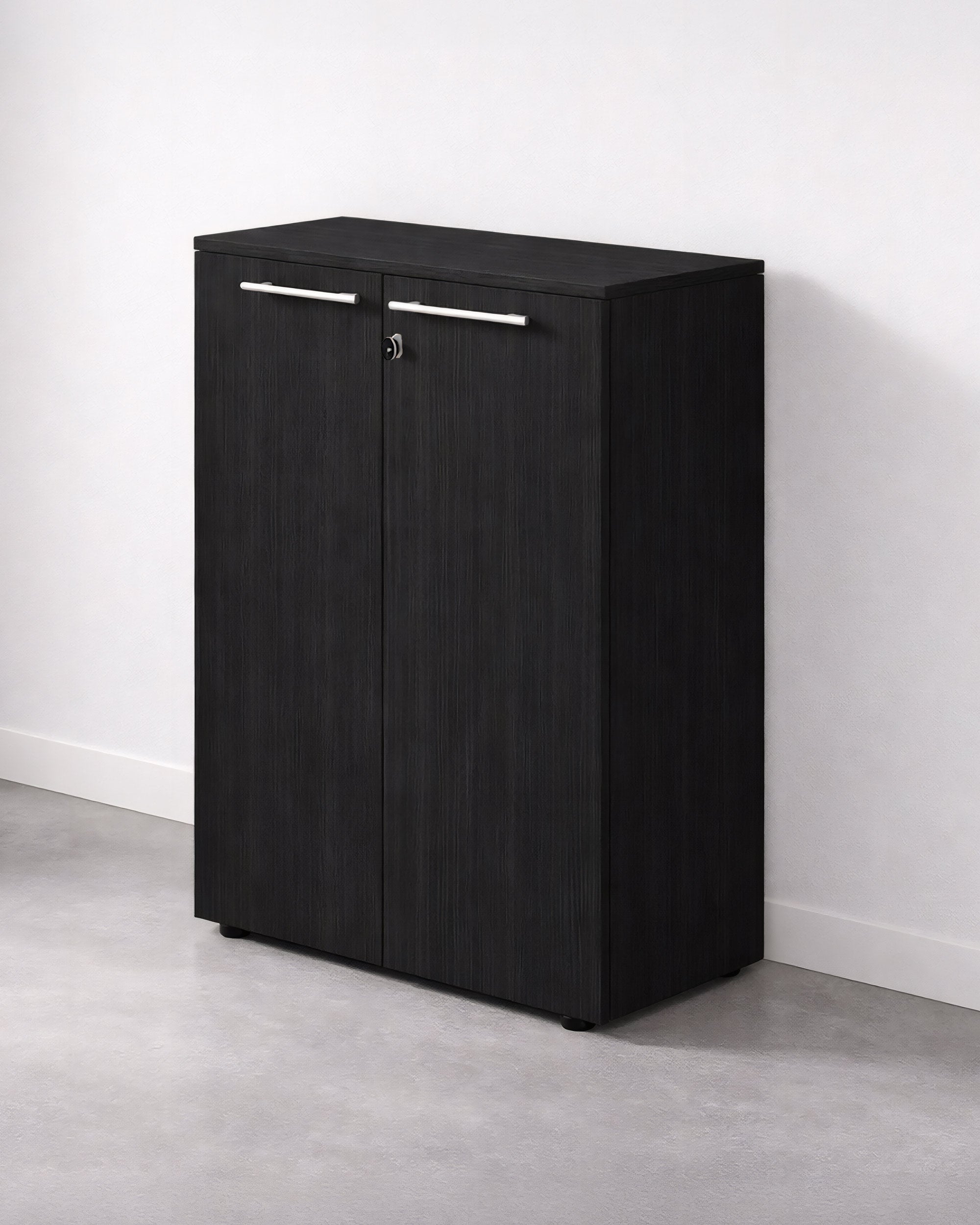 L'Armoire Mi-Haute de Bureau Flora à Portes Battantes de Monsieur Bureau, en bois noir avec deux portes, des poignées argentées et une serrure, est posée sur un sol gris contre un mur blanc - parfaite pour le rangement d'un bureau.#finitions_frene_noir