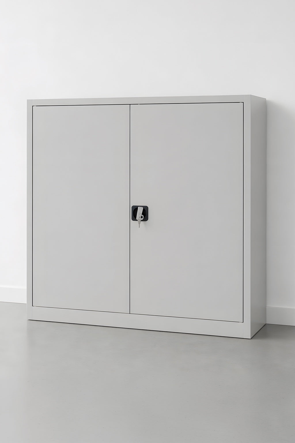 L'armoire Monsieur Bureau Basse Métal à portes battantes SOLID H100 cm est un meuble de rangement en métal gris clair avec deux portes fermées et une serrure centrale noire, offrant un rangement solide contre un mur blanc sur un sol gris.