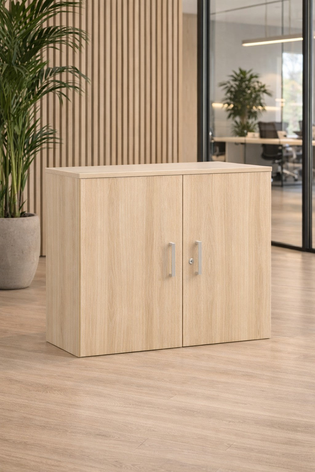 L'armoire Monsieur Bureau basse de bureau Tempo & Delta L80xH73 cm avec deux portes à poignées métalliques est posée sur un sol en bois assorti dans un bureau moderne avec des murs en verre, des plantes d'intérieur et un mobilier tertiaire élégant pour un rangement professionnel optimal.#finitions_acacia_clair