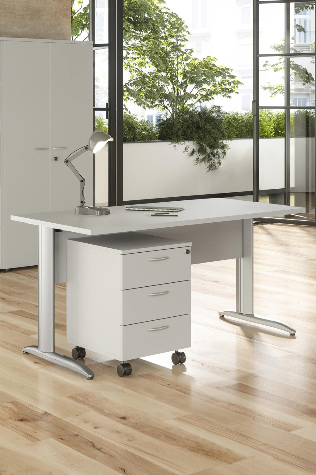 Bureau moderne avec le Bureau Pro Effect Droit ou Angle Modulable de Monsieur Bureau - bureau blanc, pieds en métal, bloc-tiroirs à roulettes ; les grandes fenêtres laissent apparaître de la verdure. Le sol et l'armoire en bois clair complètent cet espace de travail d'inspiration italienne.