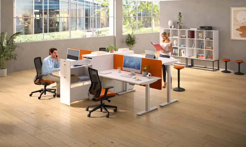 Dans un bureau moderne et lumineux, un homme utilise un Monsieur Bureau Bureau Assis-Debout Top & Down Double Moteur pendant qu'il est au téléphone et qu'une femme lit un dossier. De grandes fenêtres, des plantes et des étagères créent un espace de travail ouvert et organisé.