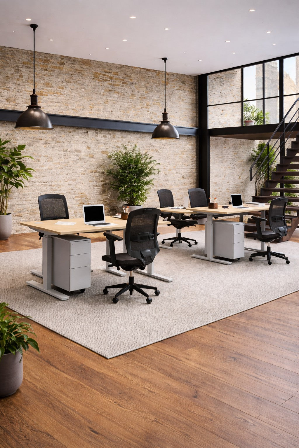 Bureau moderne à aire ouverte avec quatre bureaux Monsieur Bureau Assis-Debout Top & Down Double Moteur, des ordinateurs portables, des chaises, de grandes fenêtres, des murs en briques apparentes, des lampes suspendues, des plantes d'intérieur et un escalier en arrière-plan.