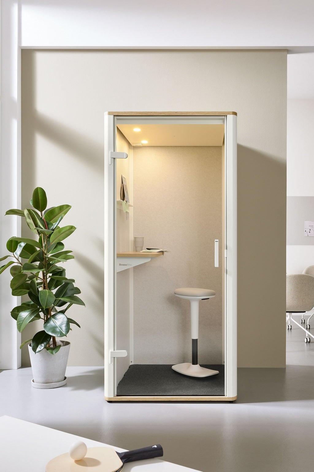 La Cabine acoustique Silen - Phone booth 1 personne de Monsieur Bureau est une cabine compacte et moderne avec un tabouret et un bureau intégré, placée contre un mur clair près d'une plante en pot dans un espace de travail lumineux et minimaliste.
