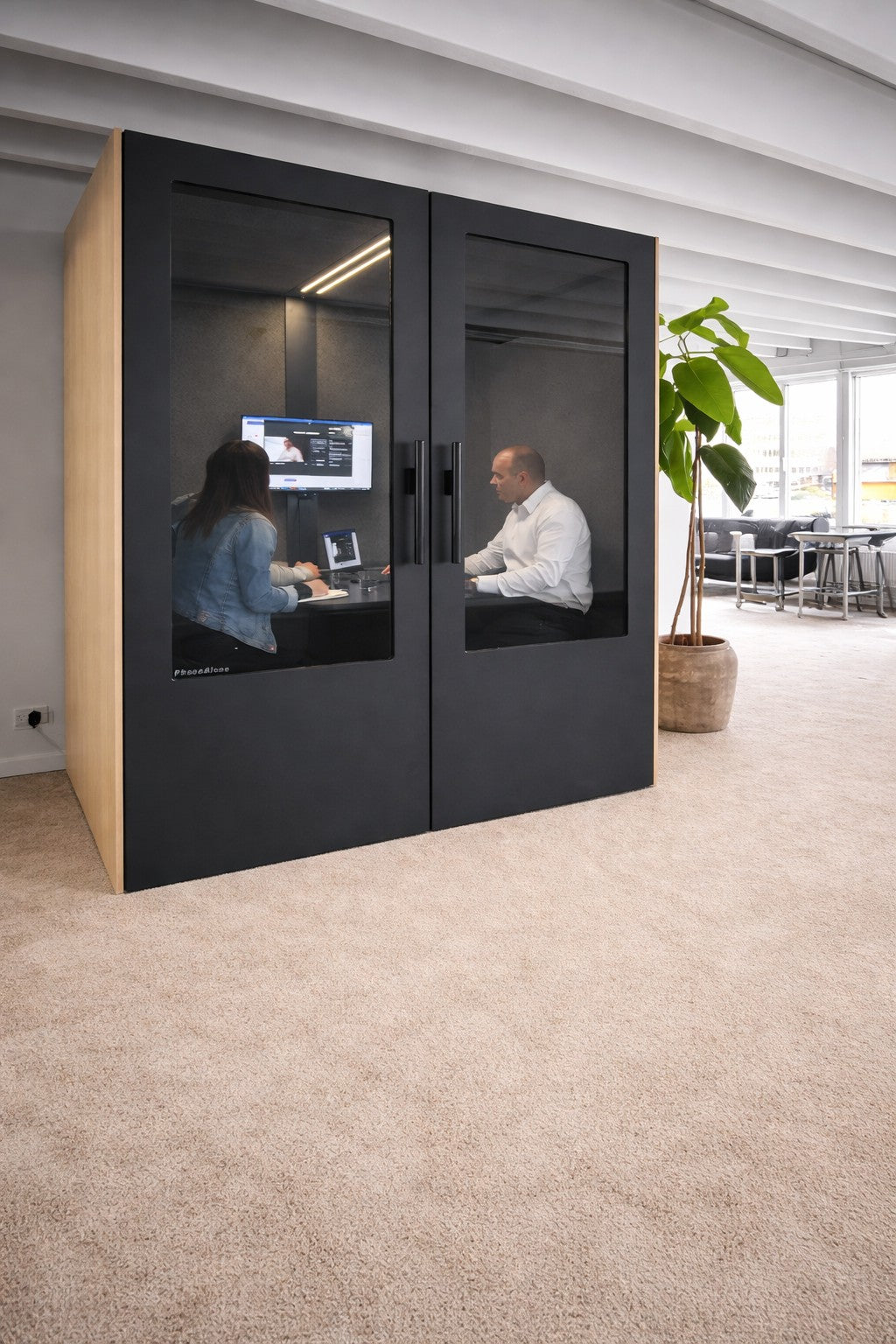 Deux personnes sont assises face à face dans un Monsieur Bureau Cabine Acoustique Réunion No Sound Standard 4 pers avec des portes en verre, travaillant sur un ordinateur ; le pod se trouve dans un bureau lumineux avec une grande plante en pot à proximité#finitions_chene