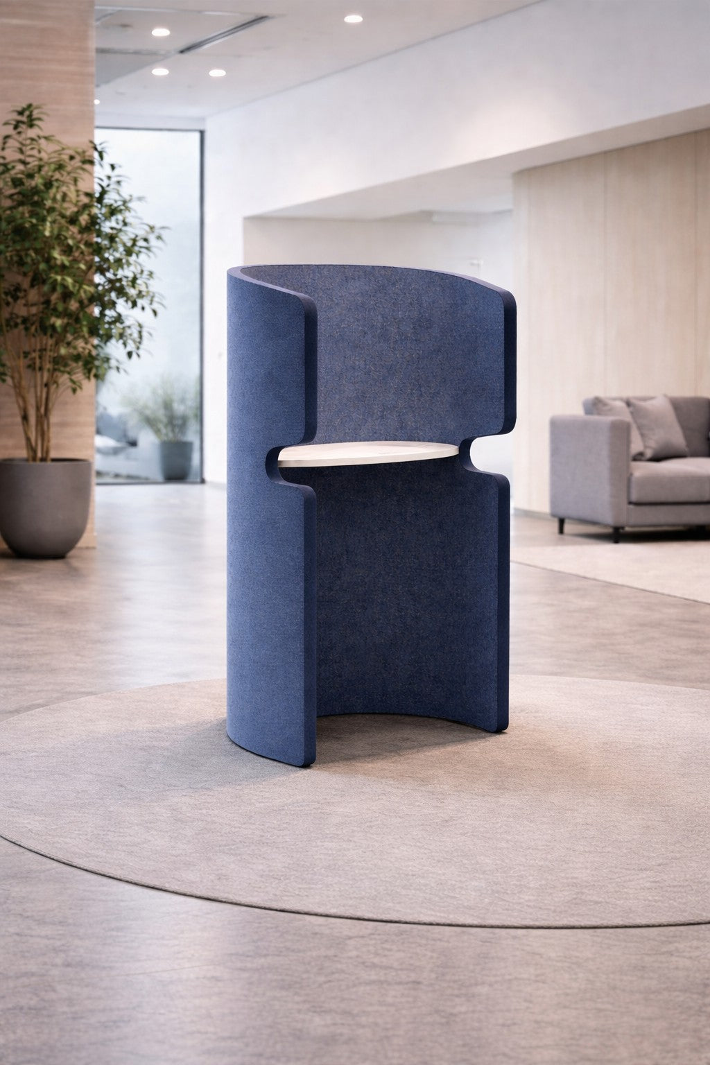 L'Alcôve acoustique debout Zen - Phone Box Open Space de Monsieur Bureau est une chaise bleue moderne à haut dossier et au design incurvé, qui offre une certaine intimité dans une pièce minimaliste aux murs clairs, au sol gris, au tapis circulaire, à la plante en pot et au canapé.