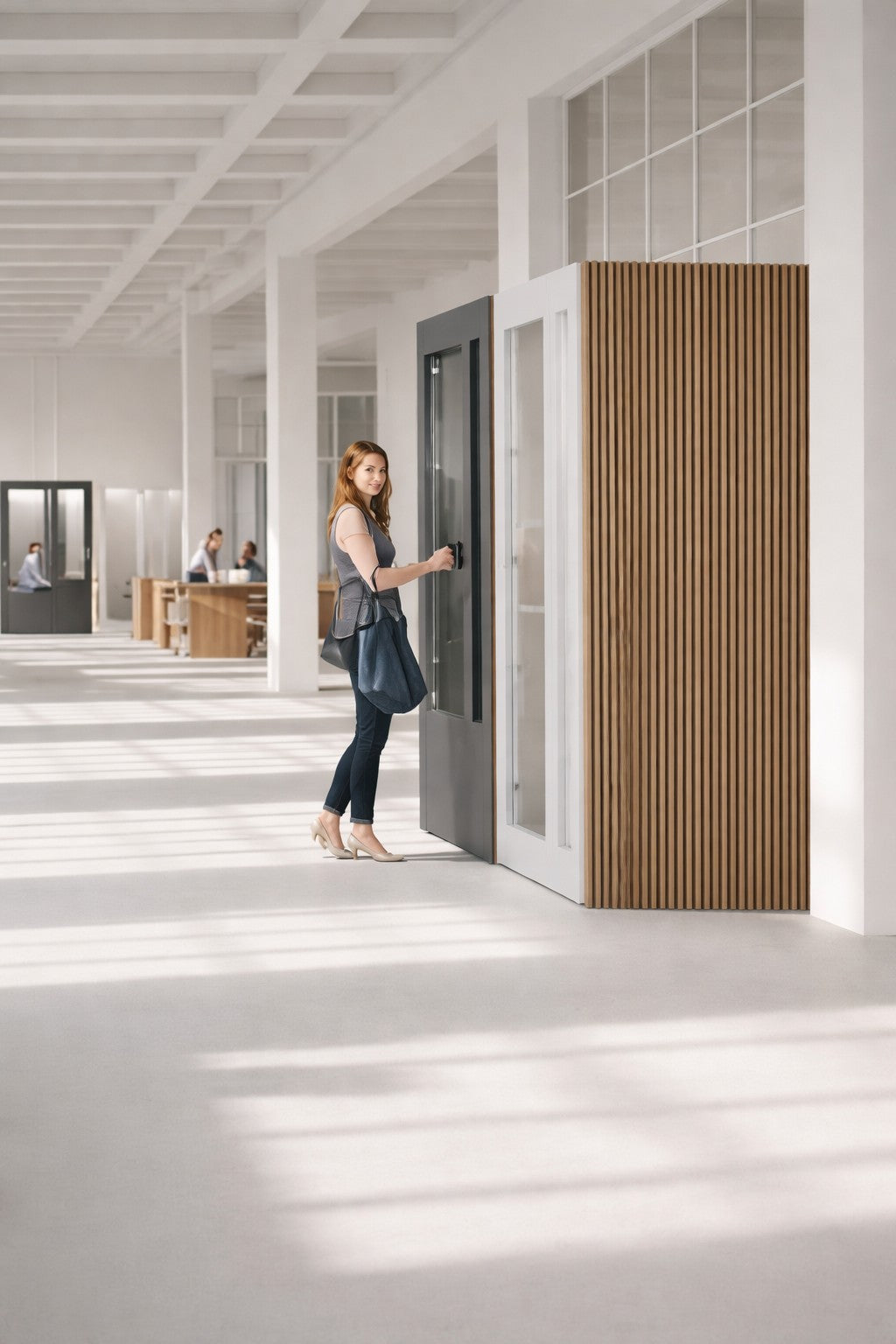Une femme aux cheveux longs, vêtue d'un haut sans manches, d'un jean et de talons, entre dans une Cabine acoustique de Travail NO SOUND Executive 1 pers de Monsieur Bureau dans un espace de travail lumineux avec de grandes fenêtres et des accents en bois pour une meilleure isolation sonore.