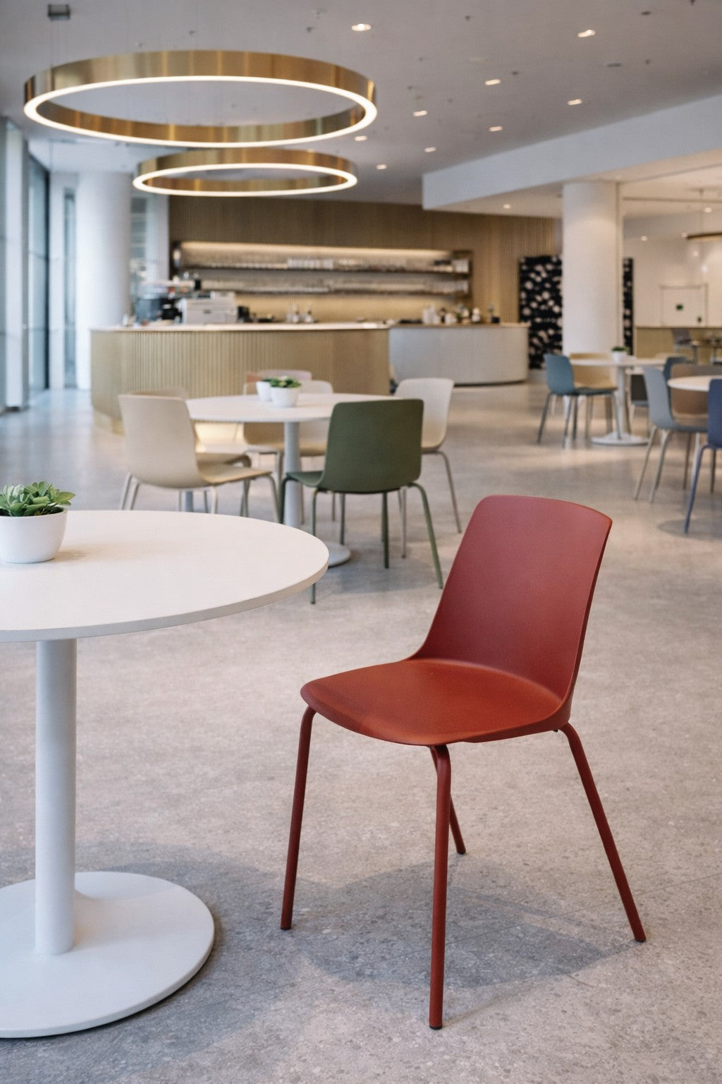 Un café moderne et spacieux avec des tables rondes, des chaises professionnelles colorées, de grands plafonniers circulaires et un comptoir minimaliste. Une petite plante est posée sur la table du premier plan à côté d'une Chaise empilable rouge ALIX de Monsieur Bureau.#finitions_rouge