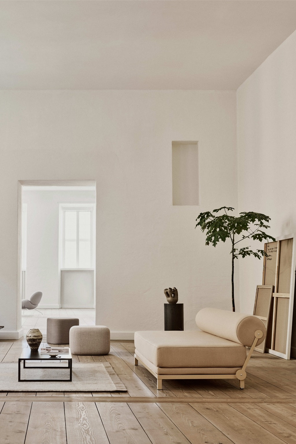 Un salon minimaliste présente le Canapé-Lit Nyx de BUSK+HERTZOG pour Monsieur Bureau, avec un sol en bois clair, une chaise longue beige, une table basse, un décor neutre, une plante en pot et des cadres vides adossés au mur.