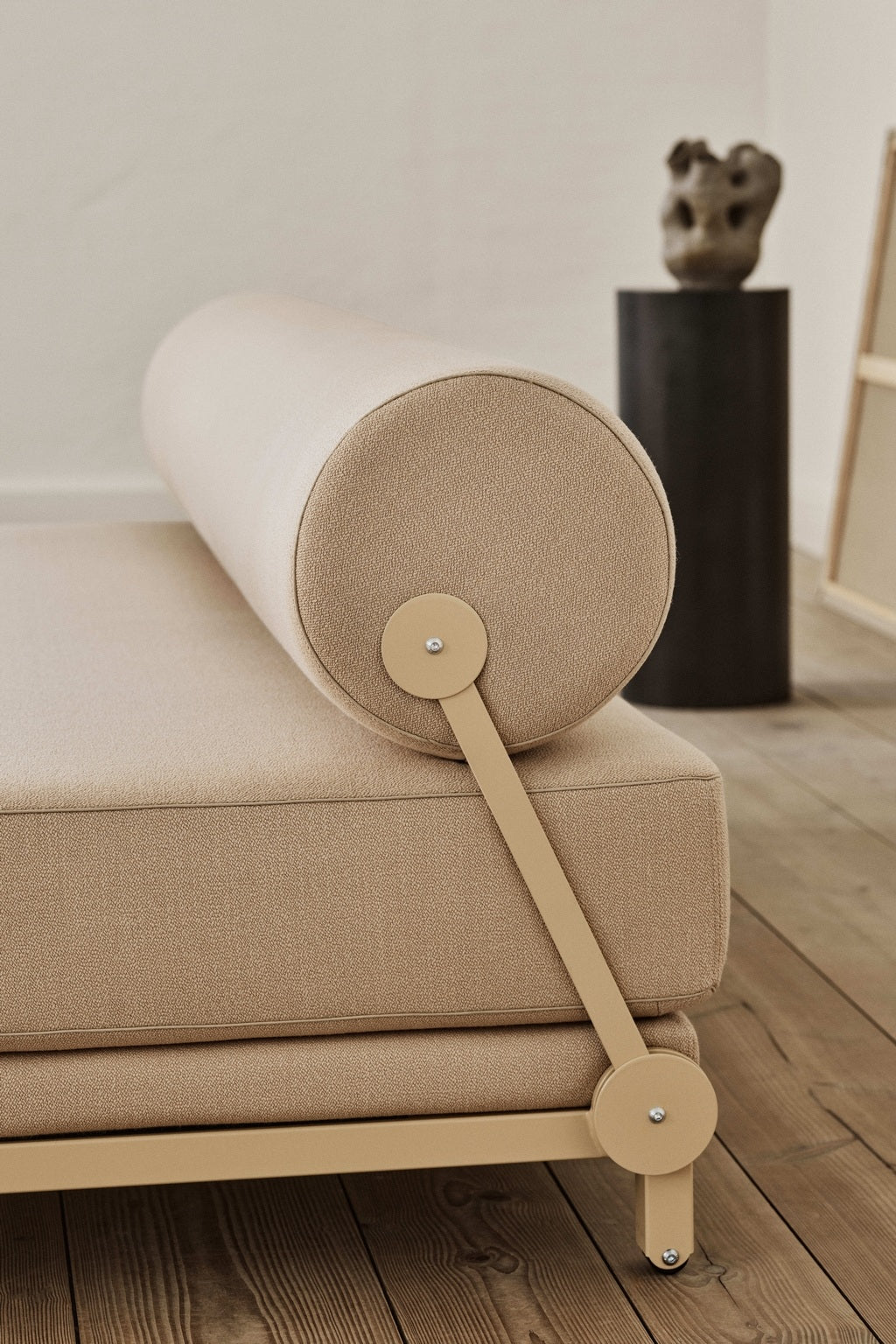 Gros plan sur le Canapé-Lit Nyx de BUSK+HERTZOG pour Monsieur Bureau, qui présente un design beige moderne avec un coussin à traversin et une structure métallique sur un sol en bois. À l'arrière-plan, un haut piédestal noir présente une sculpture abstraite.