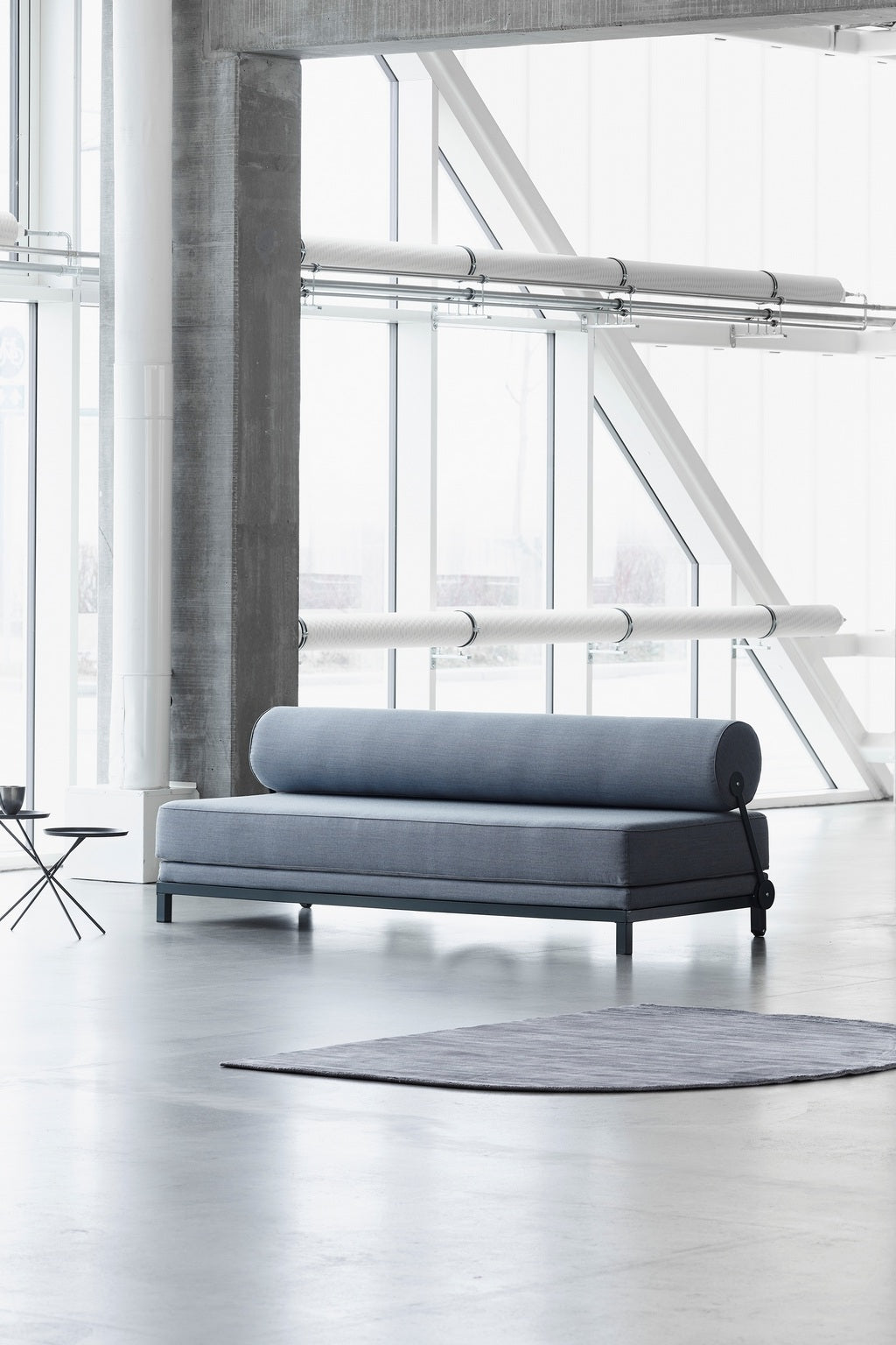 Une pièce moderne avec de grandes fenêtres, des piliers en béton, le Canapé-Lit Nyx de BUSK+HERTZOG de Monsieur Bureau en tissu bleu avec un oreiller cylindrique, une petite table d'appoint noire et un tapis gris sur un sol clair.#finitions_bleu