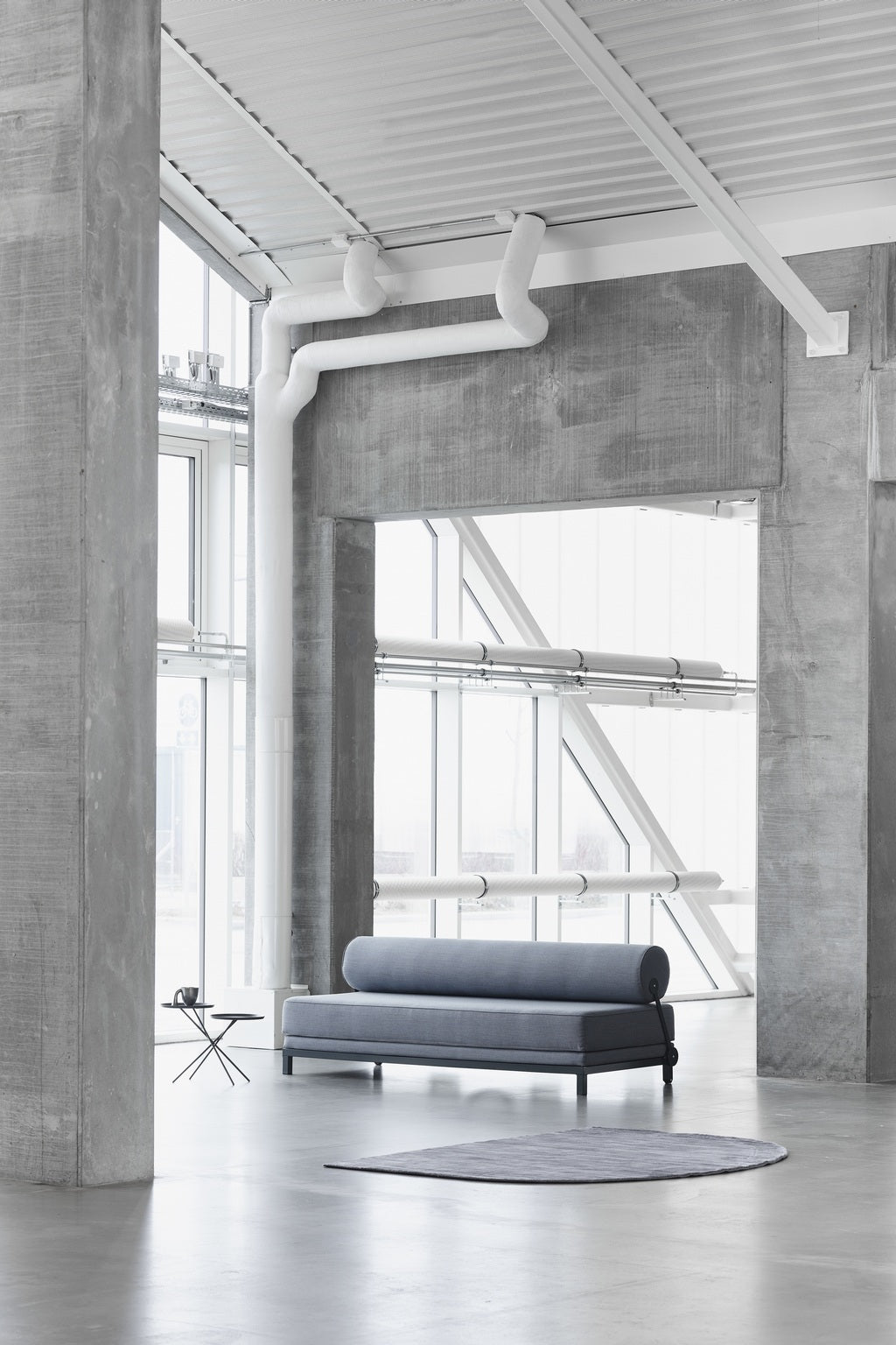 Une pièce minimaliste et moderne avec des murs et un sol en béton, de grandes fenêtres, un Canapé-Lit Nyx de BUSK+HERTZOG pour Monsieur Bureau en revêtement bleu, une petite table d'appoint ronde, un tapis rond gris et des tuyaux blancs apparents sur les murs et au plafond.