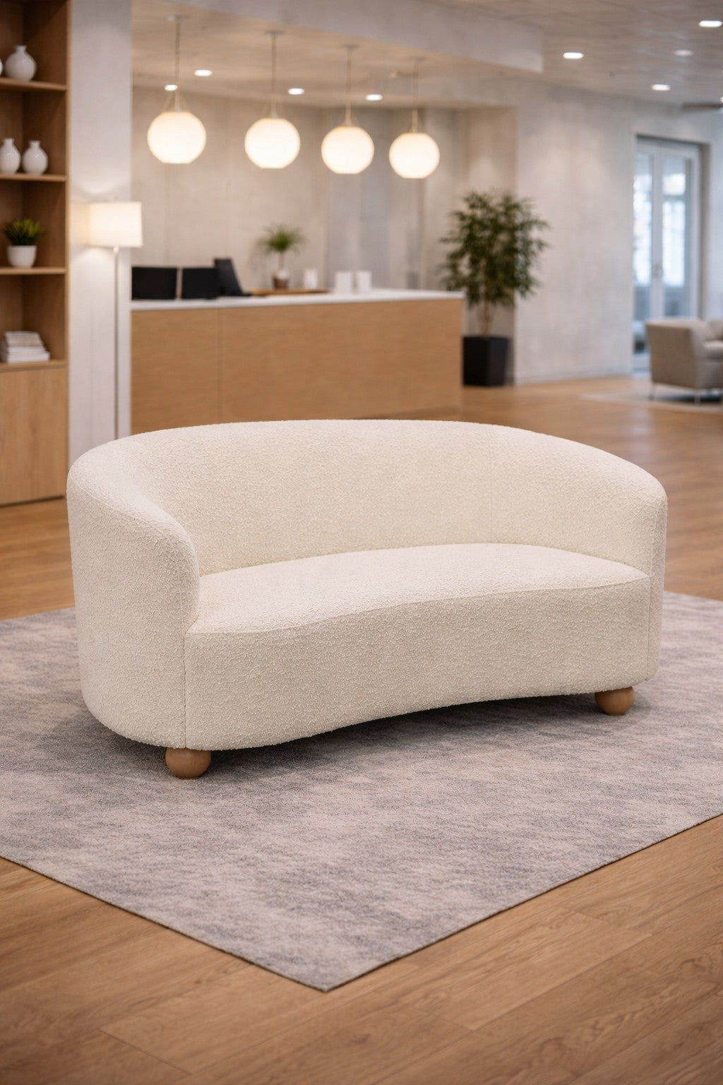 Le Canapé d'accueil Kurl 2 places tissu bouclette de Monsieur Bureau, en crème avec des pieds en bois, est posé sur un tapis gris dans une pièce spacieuse et lumineuse avec des planchers en bois, un décor minimaliste et des lampes suspendues rondes.