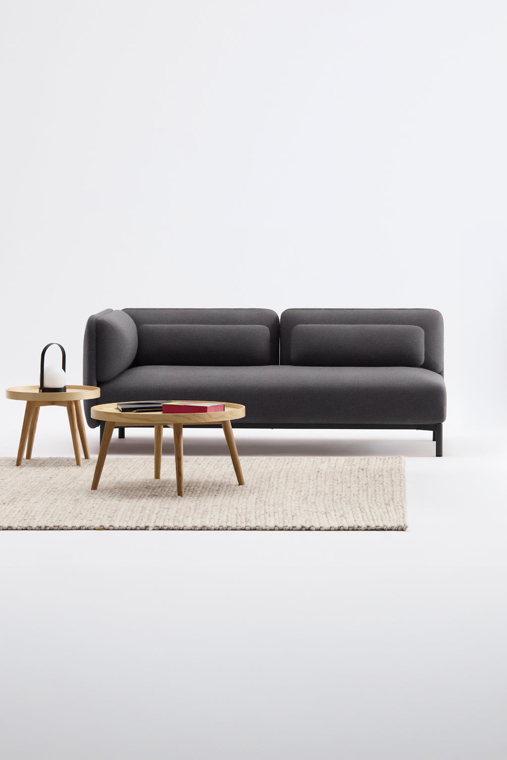 Un salon moderne présente le Canapé d'accueil modulaire Aurea - 1 accoudoir de Monsieur Bureau, associé à un canapé gris foncé, deux tables basses en bois clair, un tapis texturé beige et une lampe noire, le tout sur fond blanc.#finitions_gris