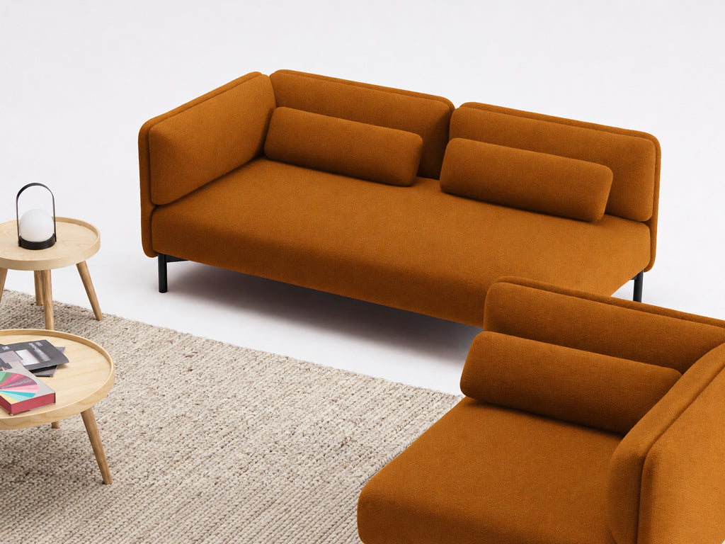 Un salon moderne présente le Canapé d'accueil modulaire Aurea - 1 accoudoir de Monsieur Bureau, deux canapés orange avec des coussins cylindriques, une table basse ronde en bois avec des livres et une lanterne, ainsi qu'un tapis beige texturé sur un sol blanc.