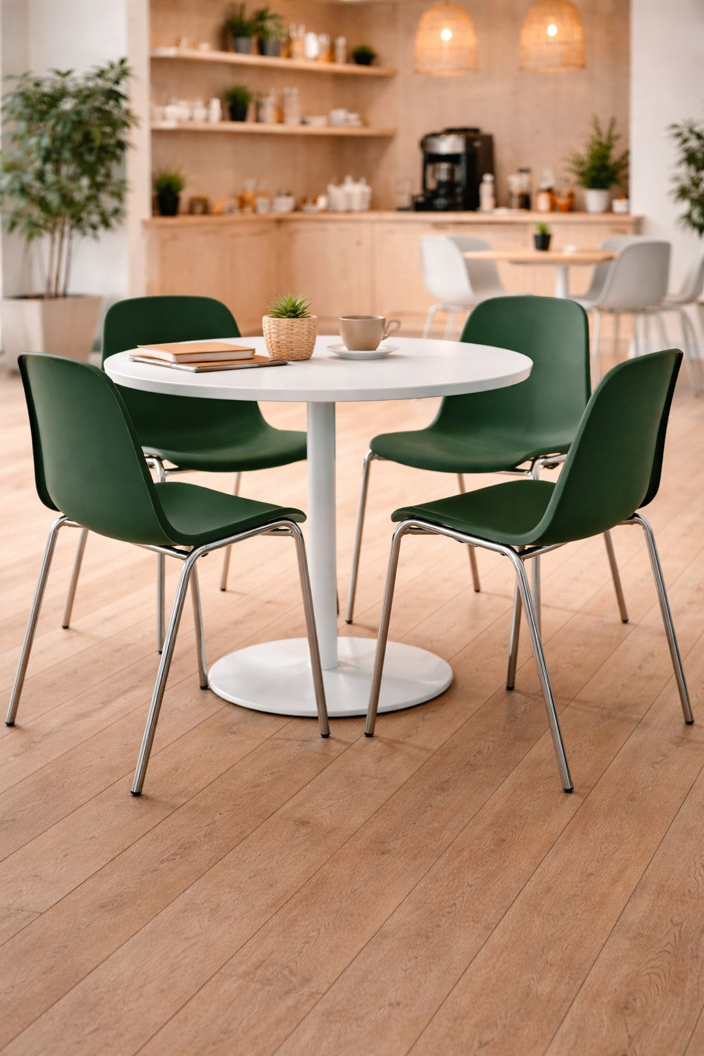Un café moderne avec une table ronde blanche entourée de quatre Monsieur Bureau Chaise ERP empilable et accrochable M2 Helena, connus pour leur résistance au feu, sur un sol en bois clair. L'arrière-plan comprend des plantes, des étagères et une station de café avec un éclairage chaleureux.#combo_vert