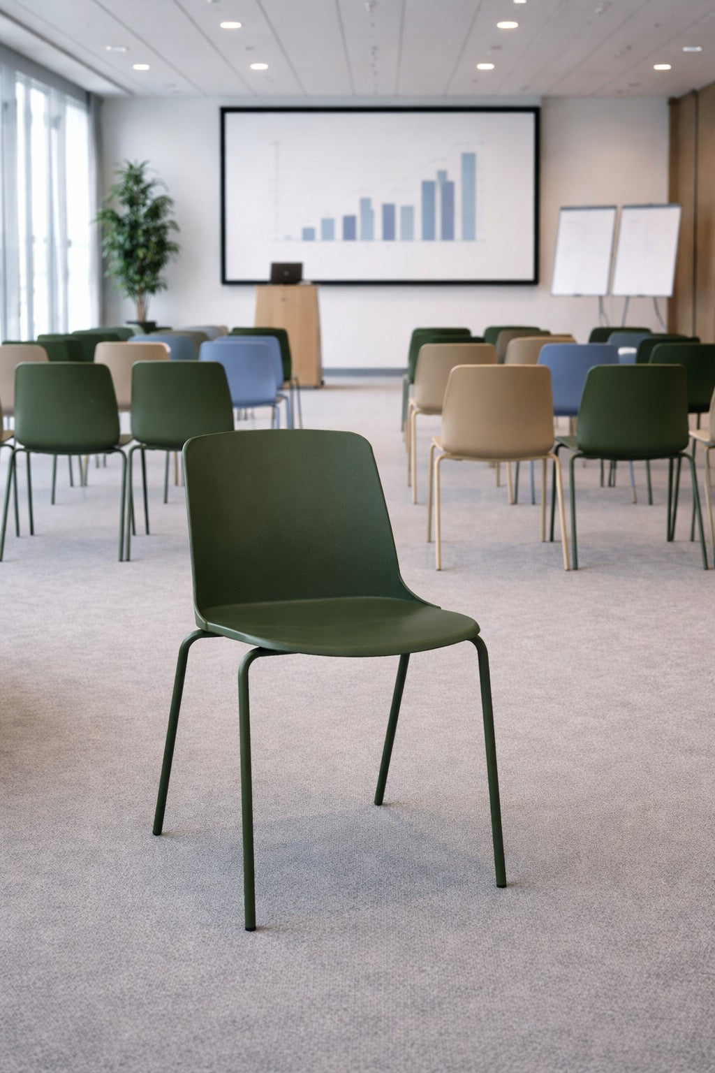 Une Chaise empilable ALIX - Polyvalente, résistante et design by Monsieur Bureau se trouve au premier plan d'une salle de conférence avec des chaises vides, un graphique à barres sur l'écran, des tableaux blancs et un podium à l'arrière-plan.#finitions_vert