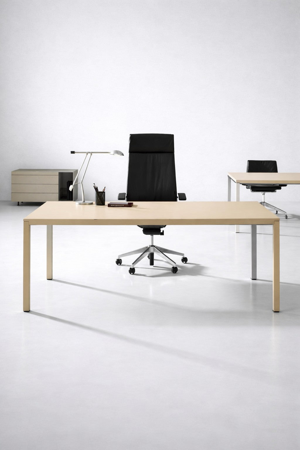 Un bureau minimaliste comprenant le Bureau de direction Nova de Monsieur Bureau (L200 x P100 cm), une chaise pivotante noire, une lampe de bureau et des fournitures de bureau. À l'arrière-plan, deux bureaux et un bloc-tiroirs sont placés contre un mur blanc.