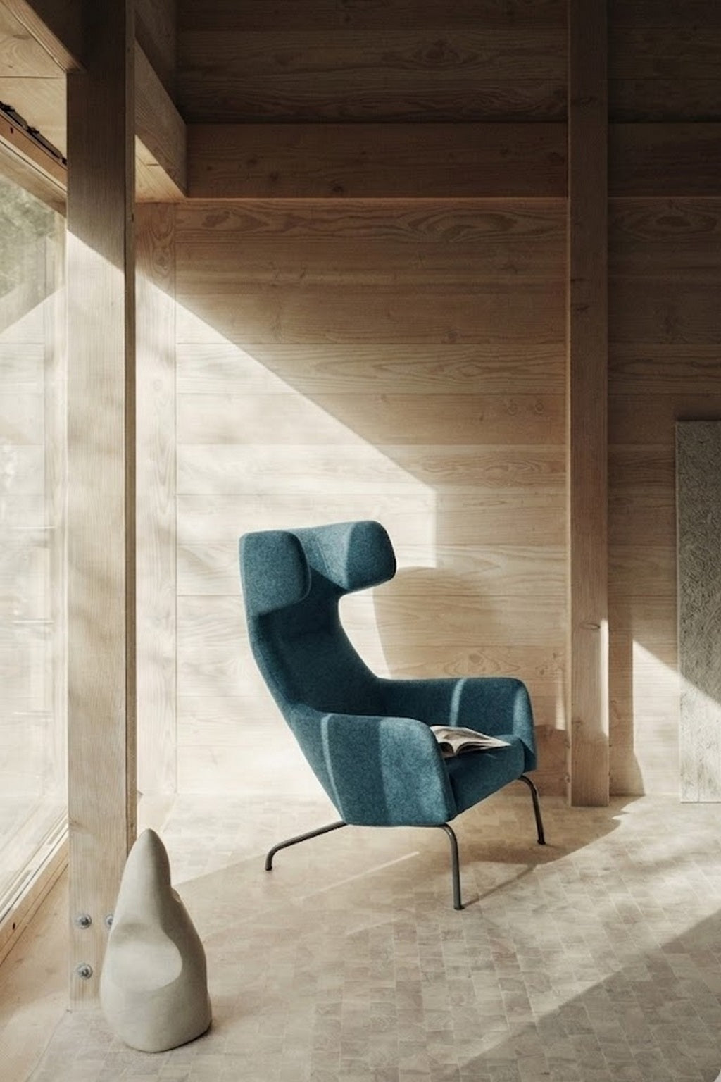 Le dossier Fauteuil lounge Fjord haut de Busk+Hertzog pour Monsieur Bureau, en sarcelle moderne, se trouve dans une pièce en bois éclairée par le soleil, avec des murs et un sol clairs. La lumière provenant d'une grande fenêtre met en valeur une sculpture abstraite et un magazine situés à proximité.#finitions_bleu_canard