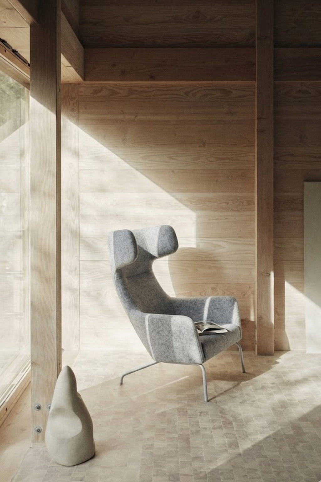 Le Fauteuil lounge Fjord haut dossier de Busk+Hertzog pour Monsieur Bureau, avec son revêtement gris moderne et ses côtés ailés, se trouve dans une pièce en bois éclairée par le soleil. Une sculpture abstraite en pierre se trouve à proximité et un magazine est posé sur la chaise.#finitions_gris_clair