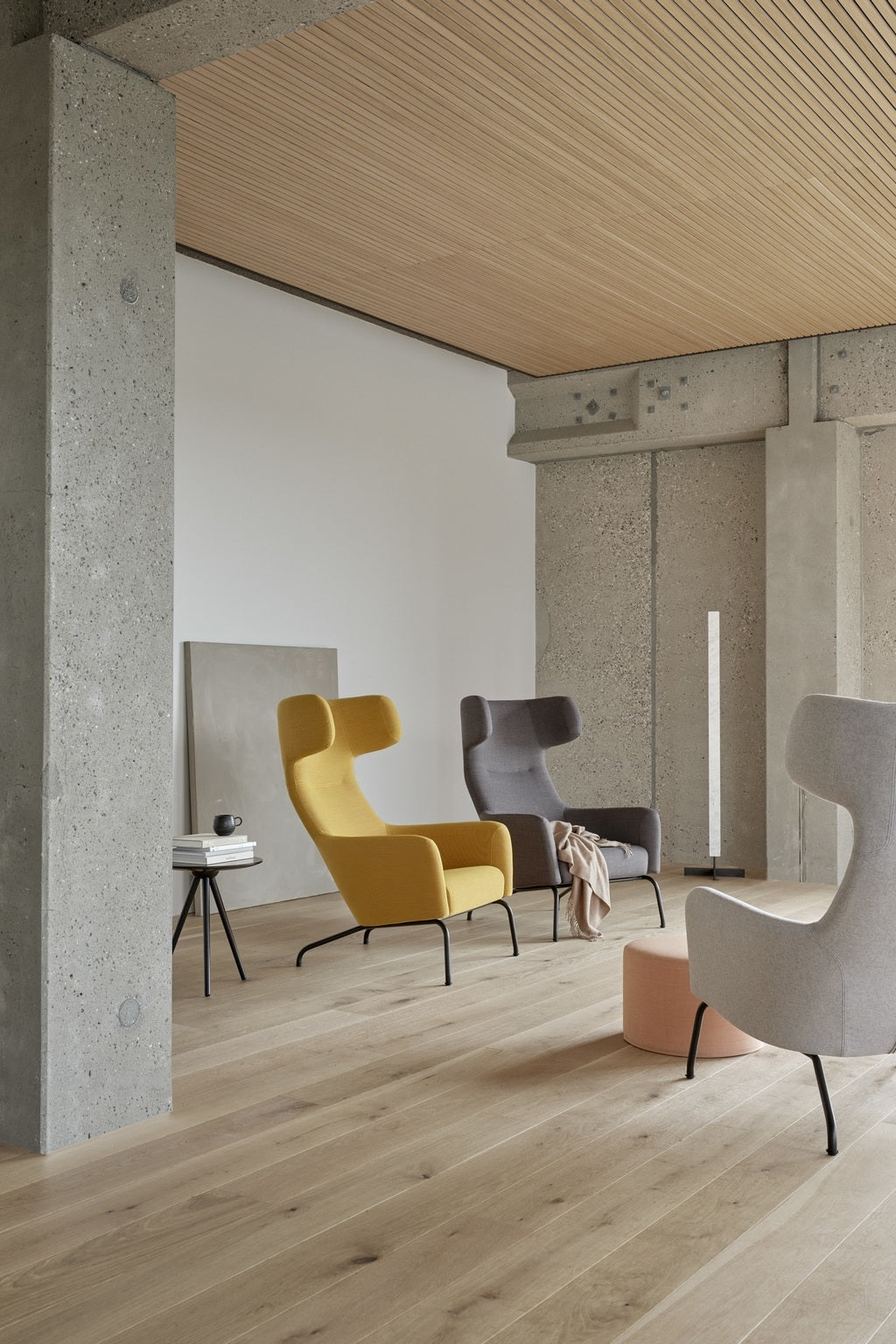 Dans une pièce moderne et minimaliste aux murs en béton et au sol en bois clair, trois fauteuils Fjord à haut dossier, créés par Busk+Hertzog pour Monsieur Bureau en jaune, gris et beige, ainsi qu'une petite table d'appoint avec un bol noir et un ottoman rose tendre, sont mis en valeur.