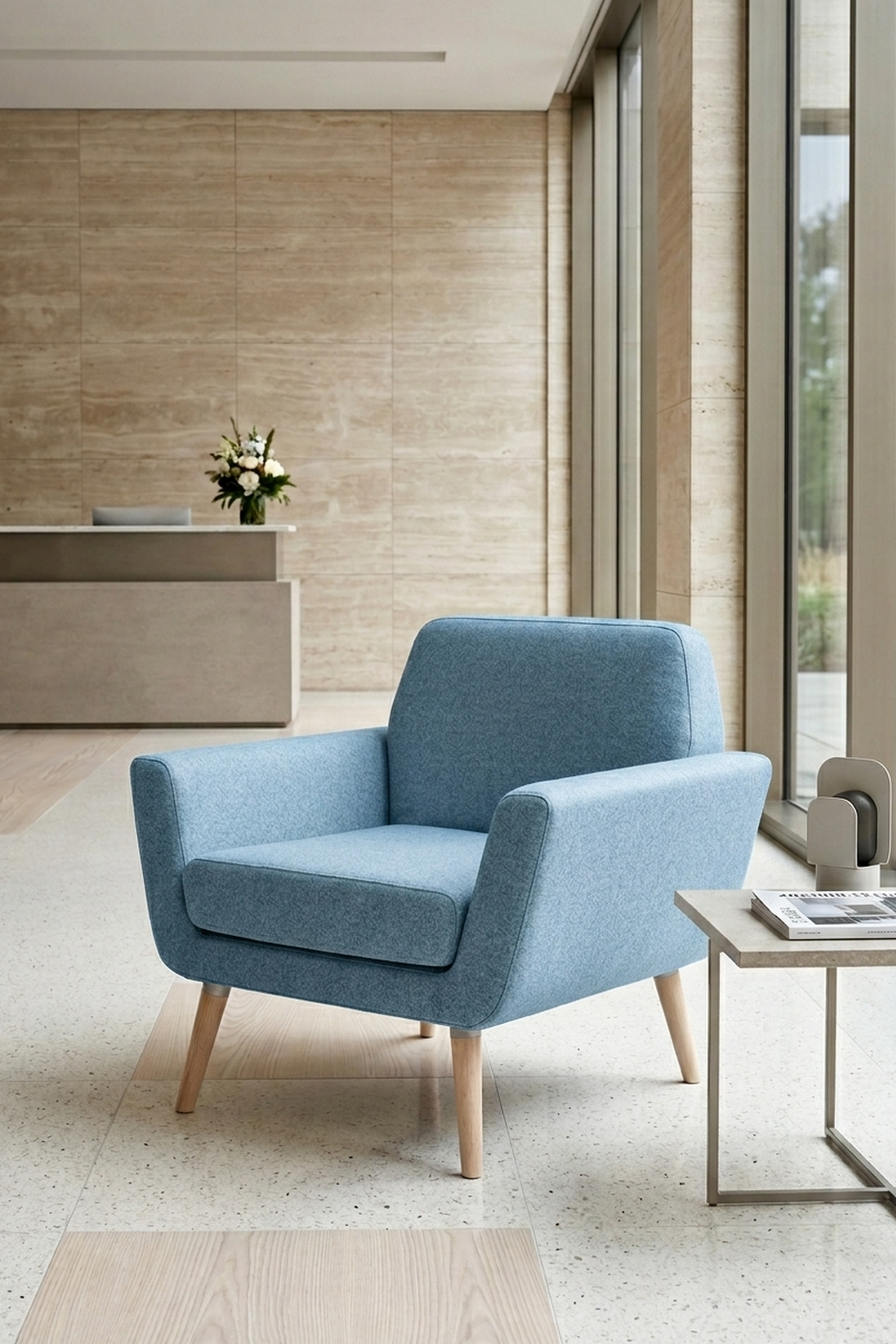 Un Fauteuil scandinave Natt de Robert ZOLLER de Monsieur Bureau, bleu clair avec des pieds en bois, est placé dans un hall d'entrée minimaliste avec une table d'appoint, des accents décoratifs, de grandes fenêtres et un bureau de réception avec des fleurs en arrière-plan.
