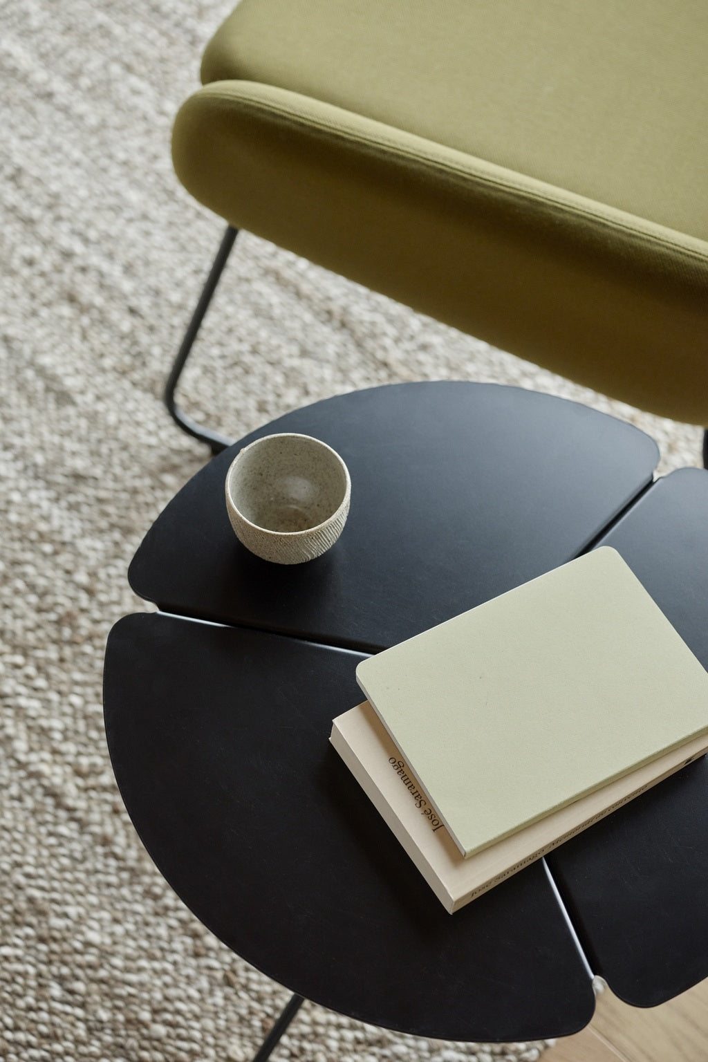 Une petite table ronde noire avec une tasse en céramique, un cahier fermé et un livre est posée à côté du Fauteuil Luge Boreale de Busk+Hertzog pour Monsieur Bureau sur un tapis tissé texturé, créant ainsi une atmosphère minimaliste et accueillante.