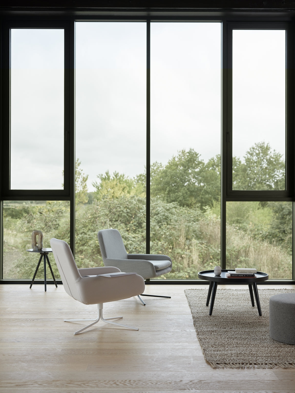 Un salon moderne avec deux chaises Fauteuil Pivotant Boreale de Monsieur Bureau (design Busk+Hertzog) et une table basse ronde sur un tapis tissé. De grandes fenêtres allant du sol au plafond encadrent une verdure luxuriante, capturant l'essence du design scandinave.