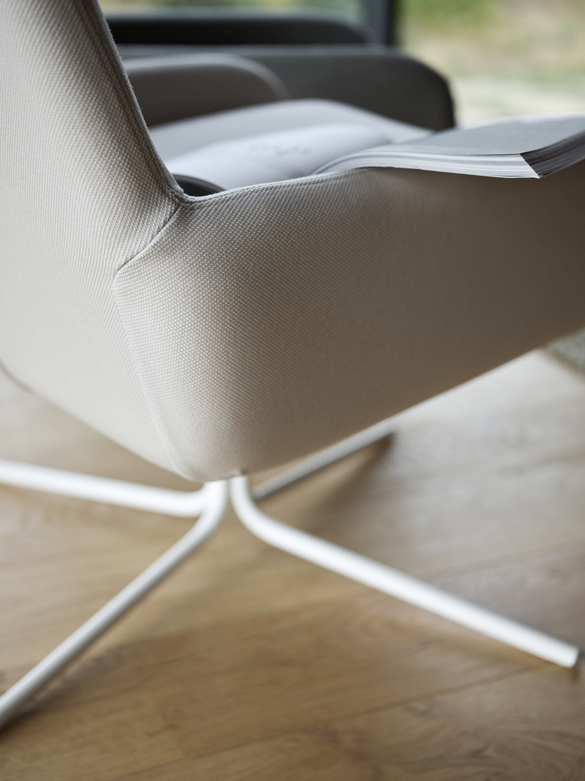 Un gros plan montre le Fauteuil Pivotant Boreale de Busk+Hertzog pour Monsieur Bureau en beige moderne avec une base pivotante en métal blanc sur un sol en bois. Un journal plié est posé sur le bras, et de la verdure est visible à travers la fenêtre située à l'arrière.