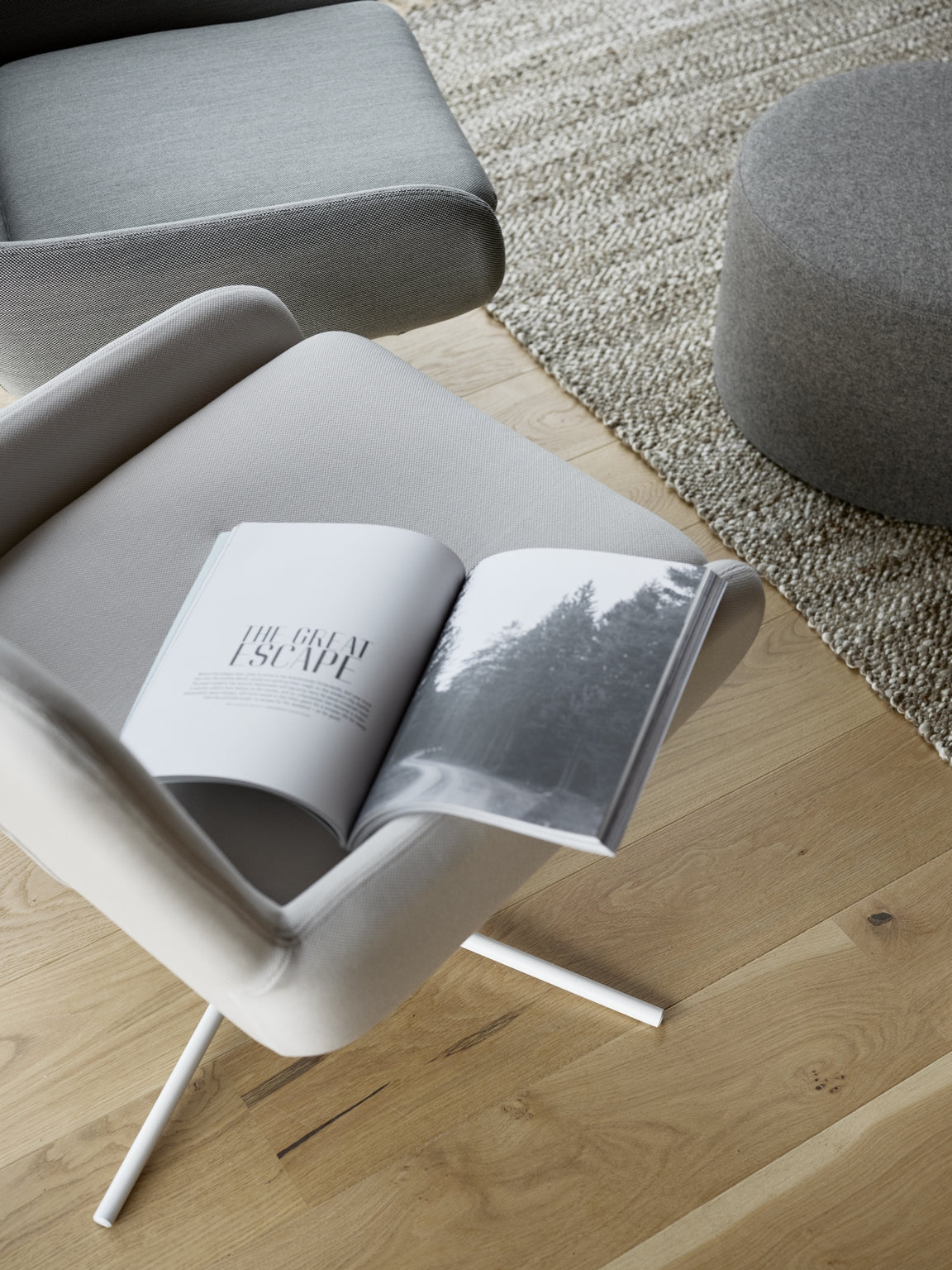 Scène minimaliste mettant en scène le Fauteuil Pivotant Boreale gris clair de Monsieur Bureau, conçu par Busk+Hertzog, tenant un magazine ouvert intitulé THE GREAT ESCAPE à côté d'un ottoman rond sur une moquette texturée et un sol en bois clair.