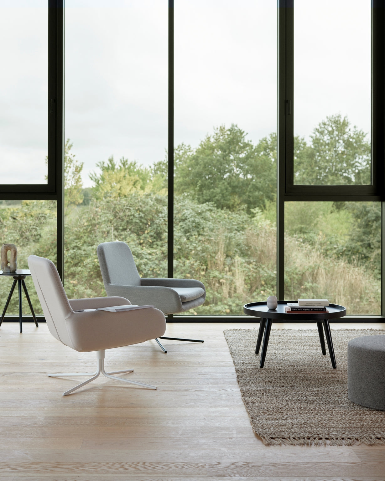 Un salon moderne au design scandinave, avec deux chaises Fauteuil Pivotant Boreale de Monsieur Bureau, une table basse ronde noire avec des livres et une tasse, un tapis tressé et de grandes fenêtres donnant sur des arbres verts.