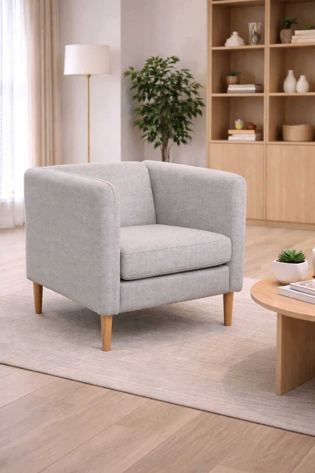 Le Fauteuil d'accueil Kanto pieds bois de Monsieur Bureau, au design scandinave et gris clair, est placé sur un tapis beige dans un salon lumineux, accompagné d'une table basse en bois avec plante et d'une bibliothèque décorée.