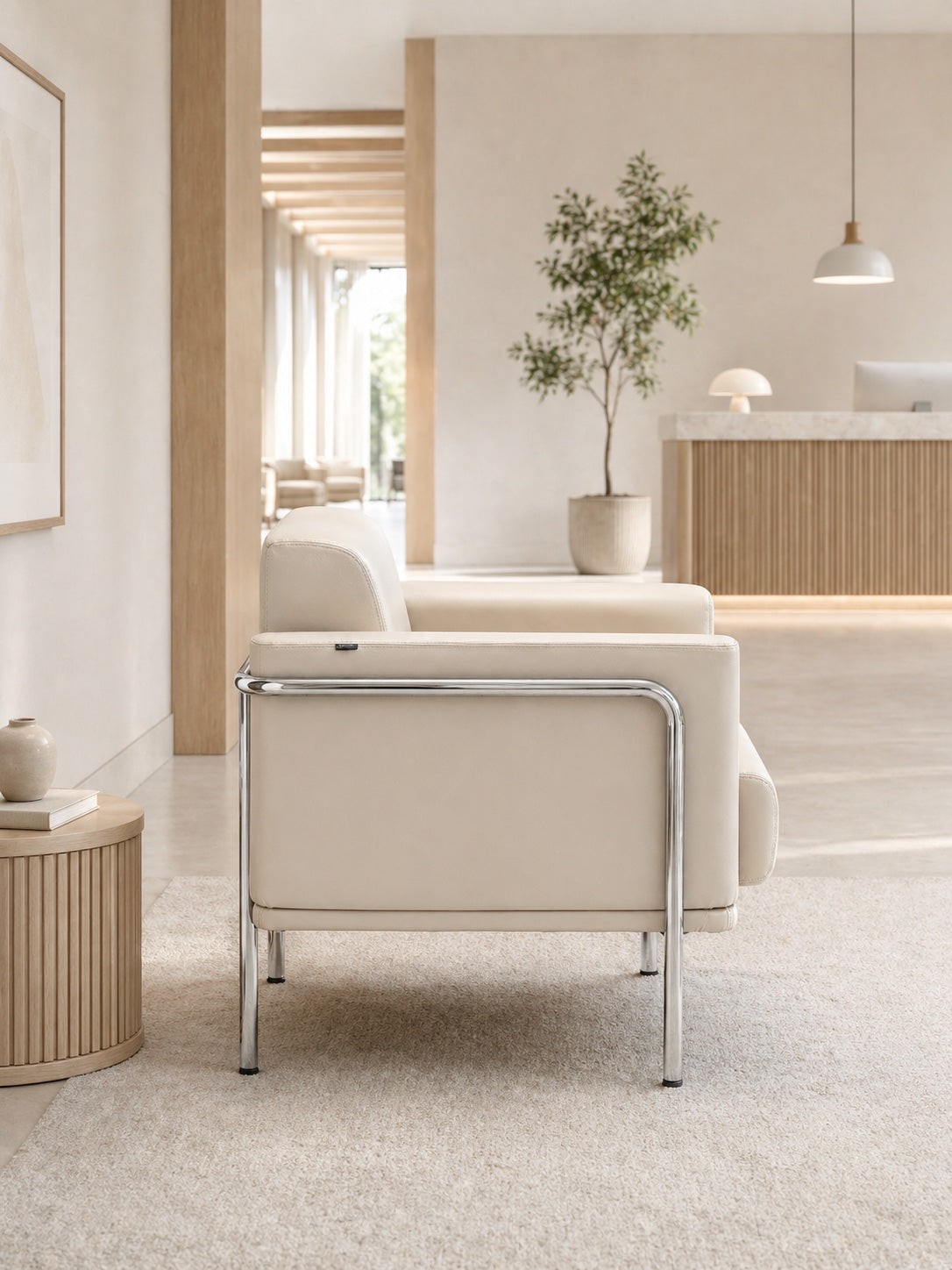 Une salle d'attente minimaliste avec le Fauteuil d'accueil Matis en simili-cuir de Monsieur Bureau aux pieds chromés, une table d'appoint et un vase, un arbre en pot, un décor neutre et la lumière naturelle créant un espace d'accueil élégant.