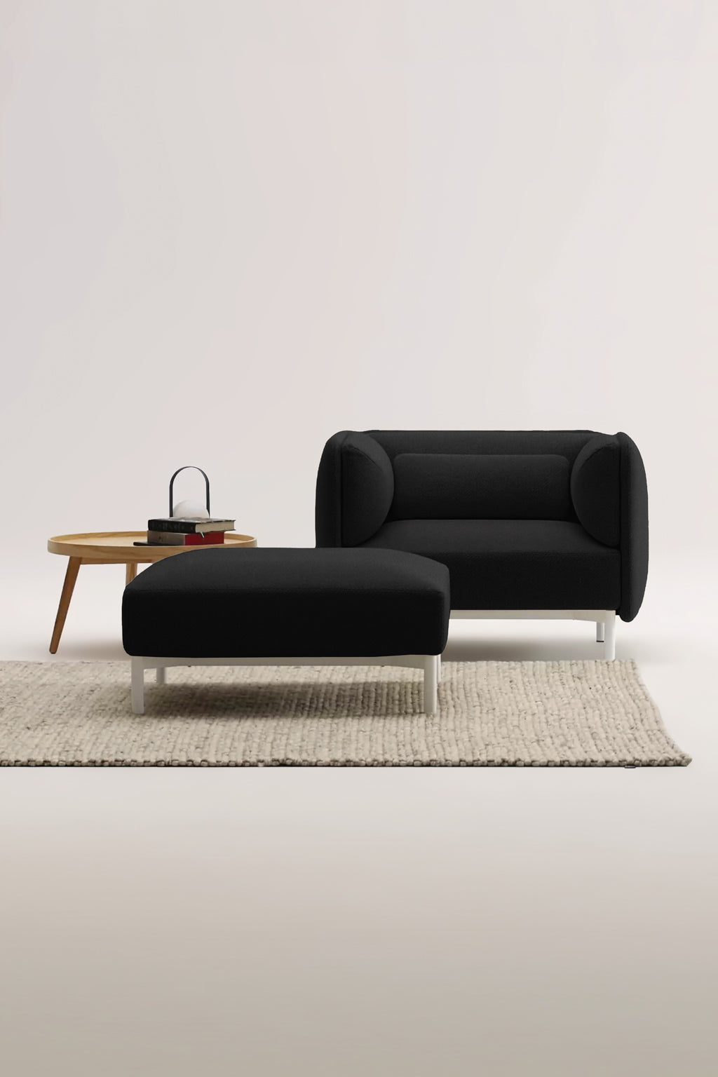 Un Fauteuil d'accueil modulaire Aurea de Monsieur Bureau, noir et moderne, avec un ottoman assorti et des pieds blancs, est posé sur un tapis beige texturé à côté d'une table d'appoint ronde en bois contenant des livres et une théière, le tout sur un fond clair et uni.#finitions_noir