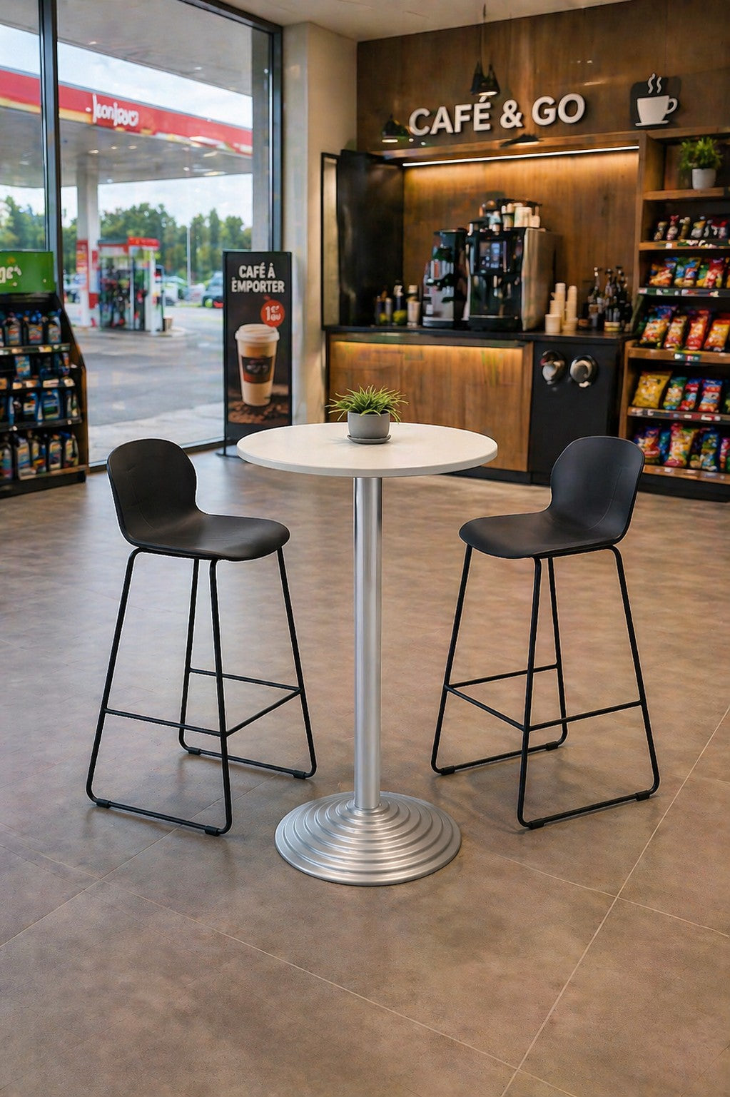 Un espace café dans une supérette avec une table ronde Monsieur Bureau Guéridon haut 6000 - Pied en Fonte, deux chaises noires, une plante en pot, des étagères de snacks, des machines à café et une grande fenêtre donnant sur la station-service.