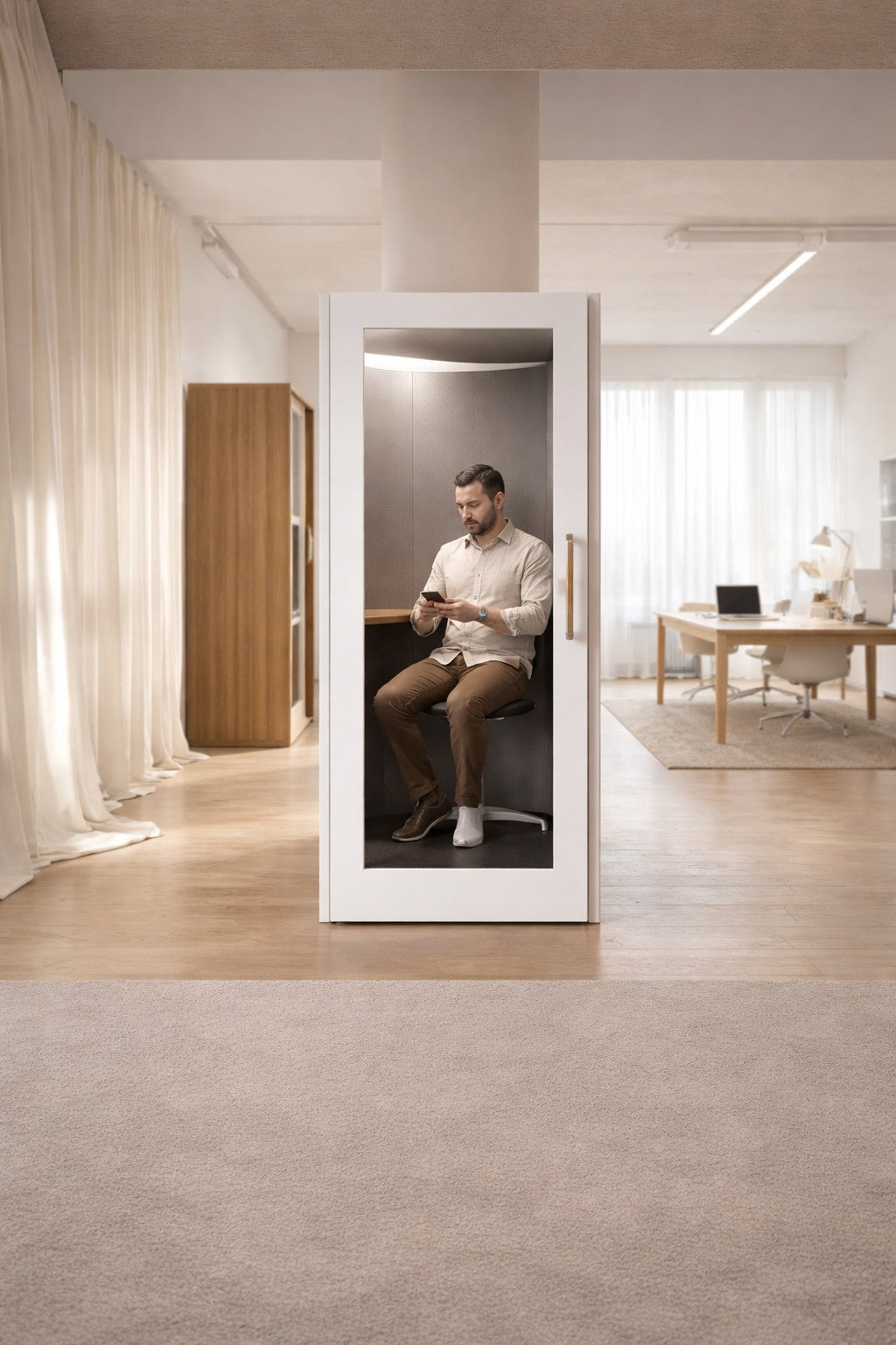 Un homme est assis à l'intérieur d'une cabine téléphonique Monsieur Bureau acoustique NO SOUND Standard 1 personne dans un bureau lumineux avec des planchers en bois et de grandes fenêtres, bénéficiant d'un isolement phonique pour un travail concentré au milieu d'un ameublement minimaliste.#finitions_blanc