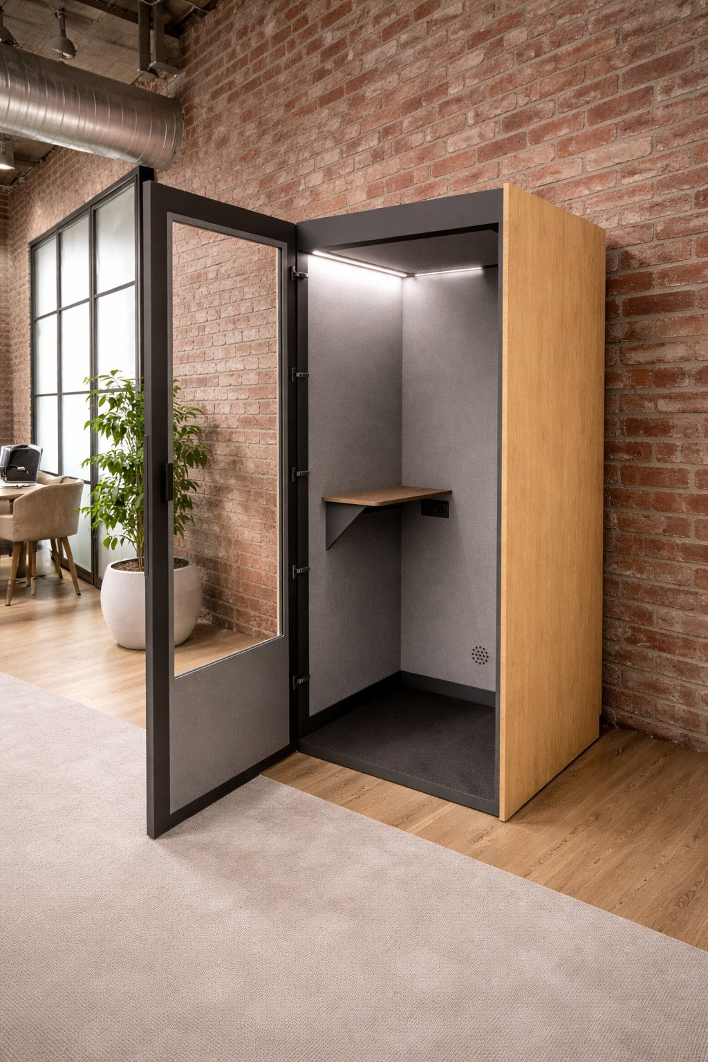 La cabine téléphonique acoustique NO SOUND Standard 1 personne de Monsieur Bureau est une cabine de bureau moderne avec un extérieur en bois, une porte en verre et une étagère, offrant un isolement phonique dans les espaces ouverts avec des murs en briques, des plantes et des bureaux.#finitions_chene