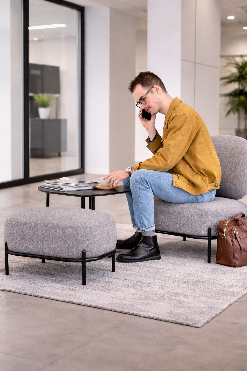 Un homme vêtu d'une veste moutarde et d'un jean est assis dans un salon moderne, parlant au téléphone tout en examinant des documents posés sur une table basse. À proximité se trouve le Pouf d'accueil Brooklin - Pieds métal de Monsieur Bureau, à côté duquel est posé un sac en cuir marron.