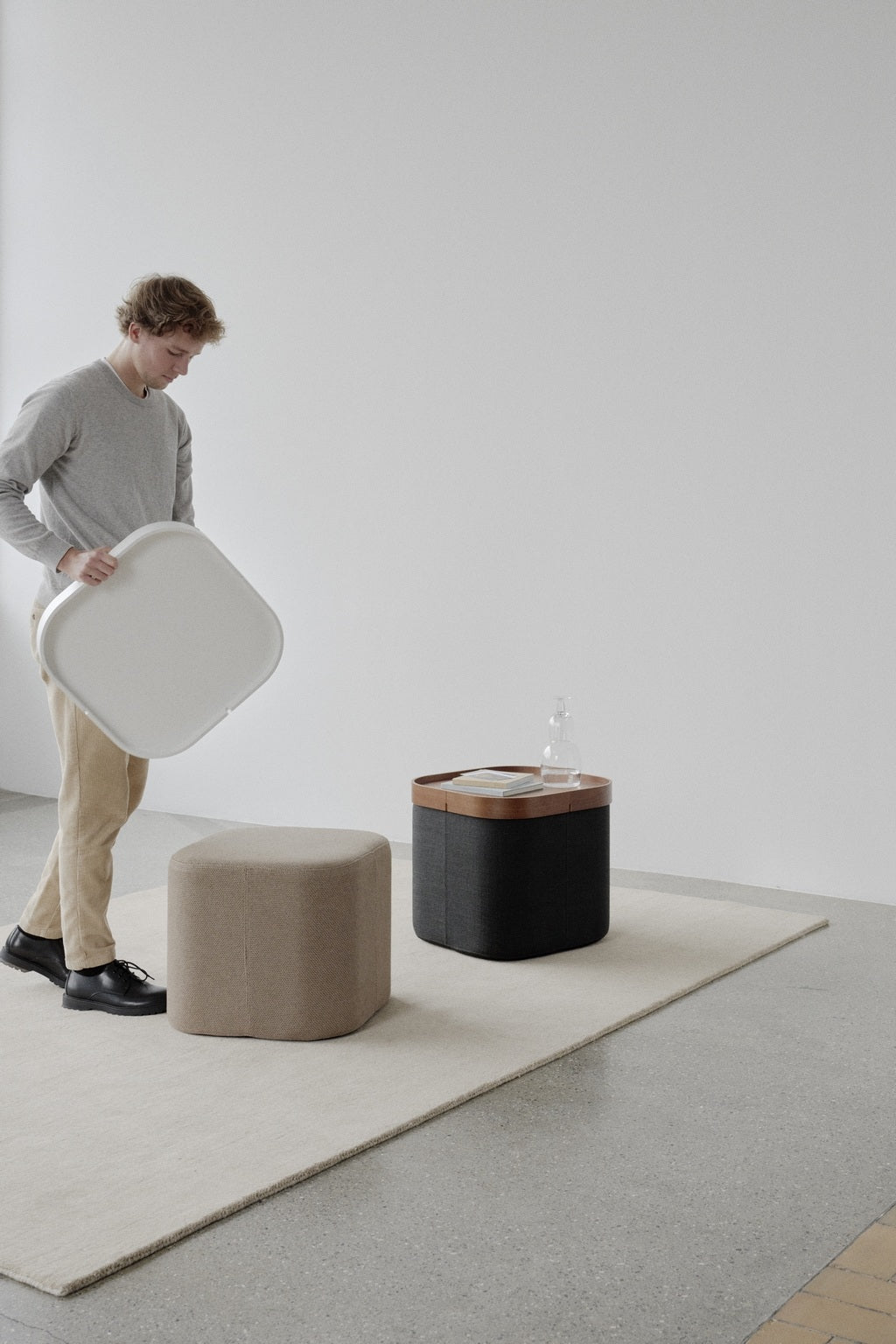 Dans une pièce minimaliste, une personne se tient debout sur un tapis clair et tient un objet blanc de forme carrée. À proximité se trouvent un Monsieur Bureau Pouf d'accueil Nordic avec plateau de Busk+Hertzog, une boîte de rangement noire avec couvercle en bois, une bouteille en verre et un livre.