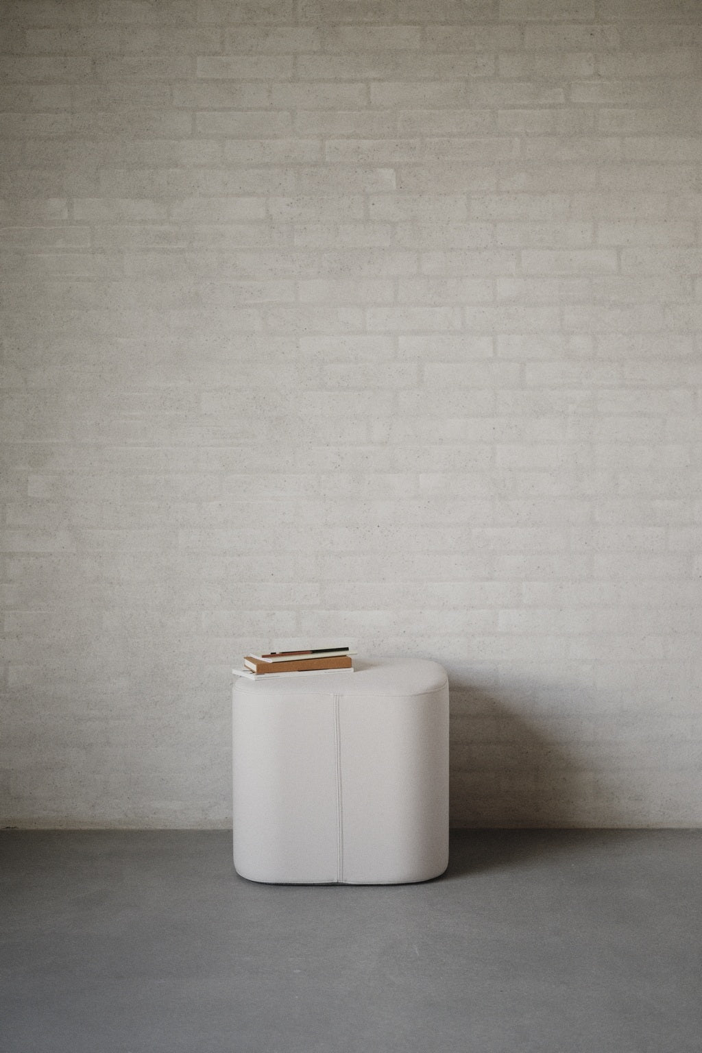 Un petit Pouf d'accueil Nordic avec plateau de Monsieur Bureau (design Busk+Hertzog) est posé sur un sol gris contre un mur de briques claires, avec deux livres fermés posés dessus.