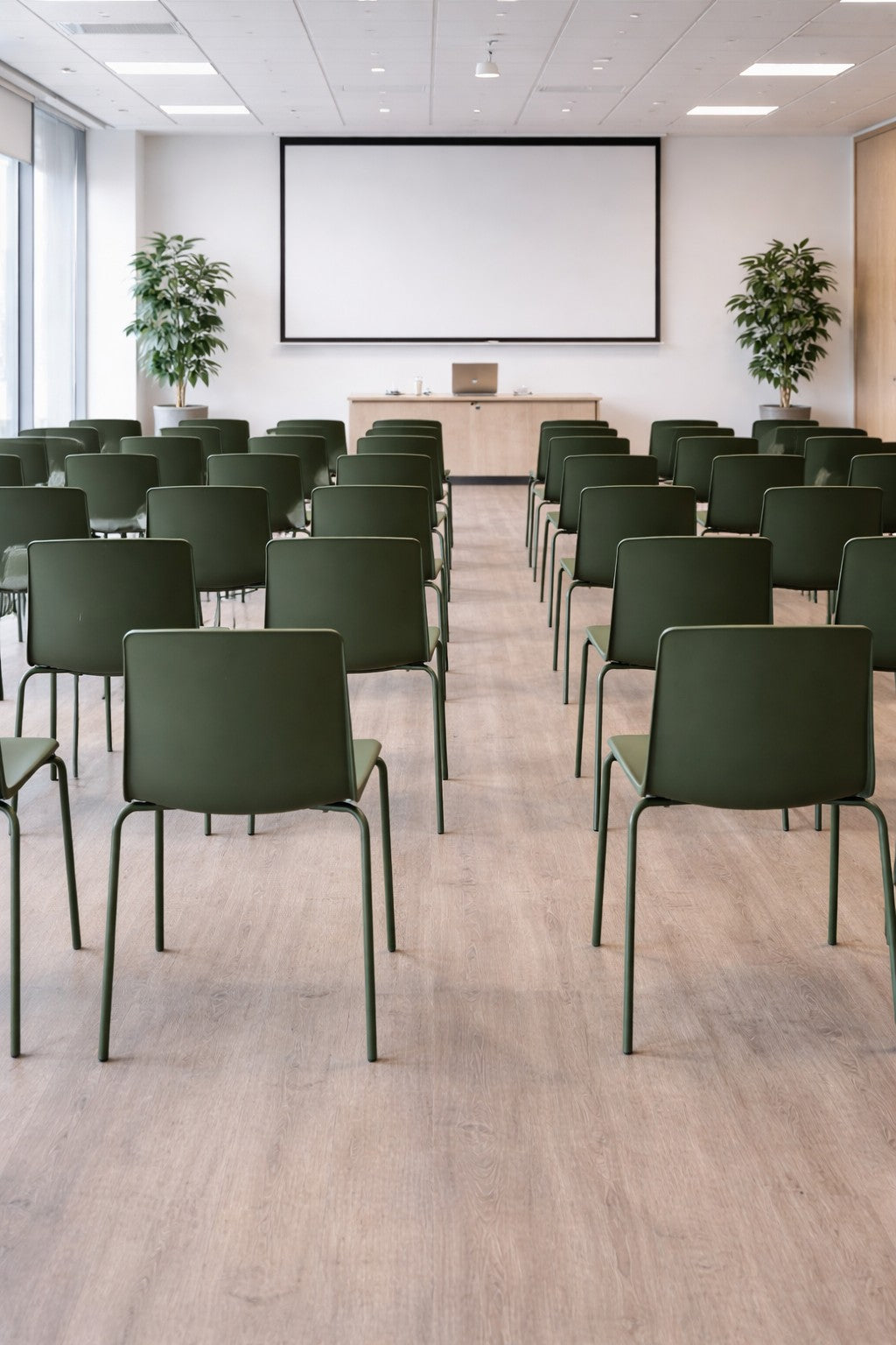Des rangées de Chaise empilable ALIX de Monsieur Bureau sont disposées dans une salle de conférence moderne avec de grandes fenêtres, un sol en bois, deux plantes en pot et une table à l'avant-polyvalente, résistante et design mobilier professionnel.