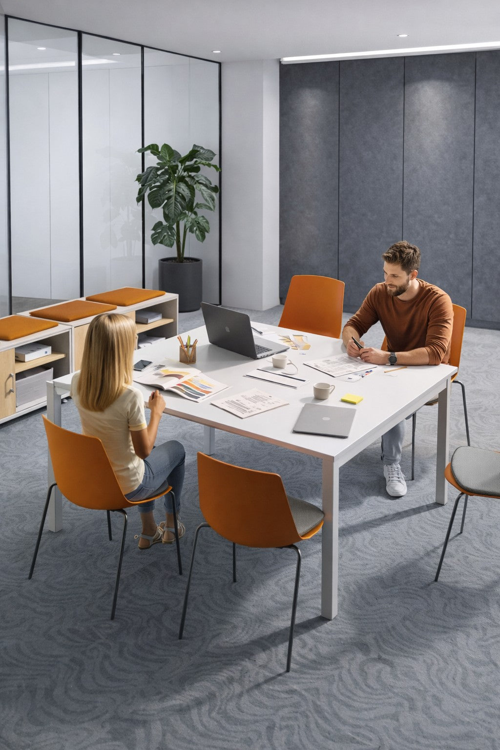 Deux personnes travaillent sur un Monsieur Bureau Solution de Table et extensions de Réunion Astral 140x143 dans une salle de réunion moderne, avec des chaises orange, une plante en pot et des dossiers sur des étagères qui améliorent l'espace de travail collaboratif.