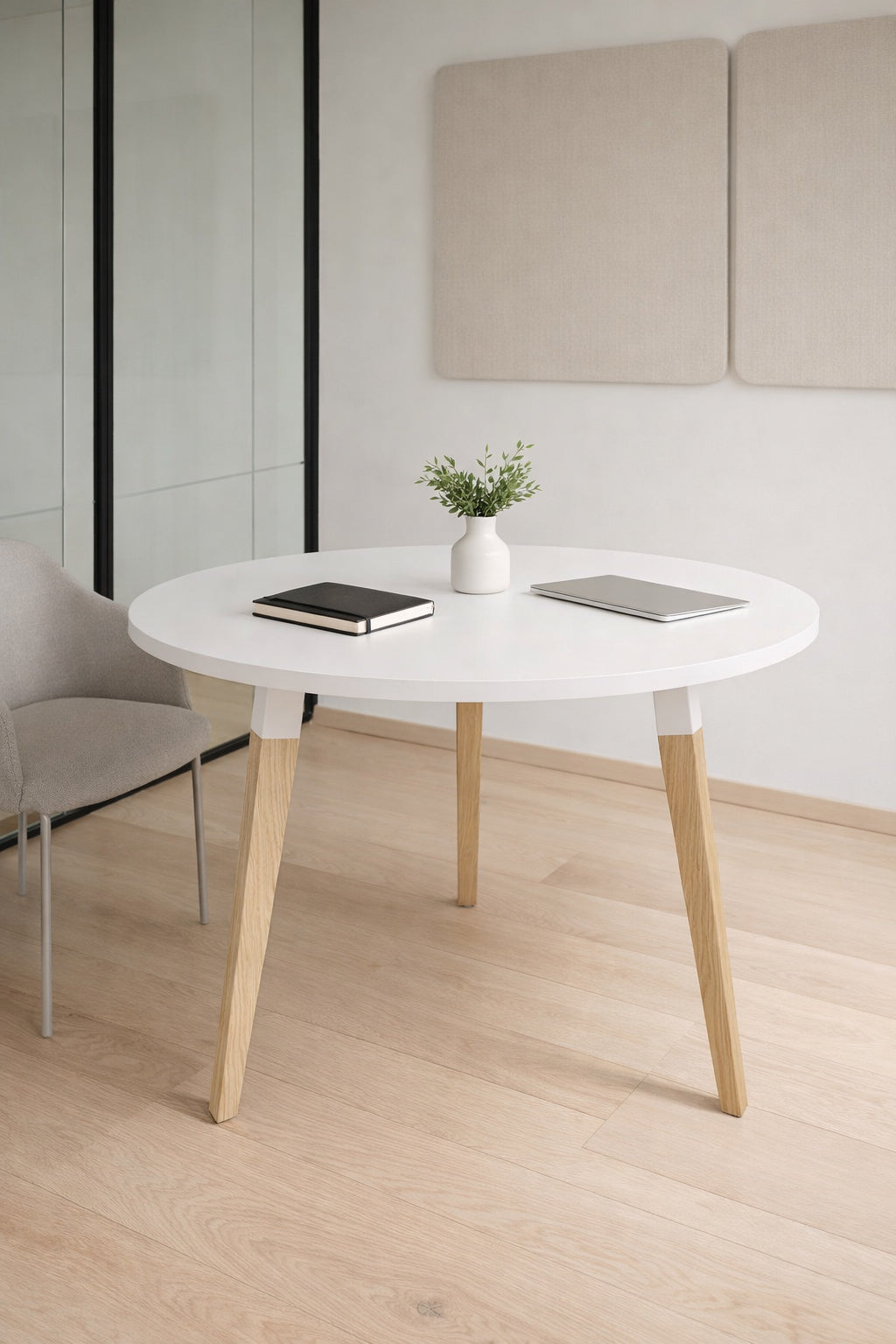 Un bureau moderne et minimal avec la Table Réunion Ronde Oslo 2 à 6 pers Ø80 à 120 de Monsieur Bureau, des pieds en bois clair, une plante en pot, un ordinateur portable fermé, un bloc-notes, une chaise rembourrée et des panneaux muraux acoustiques - parfaits pour les espaces de coworking.