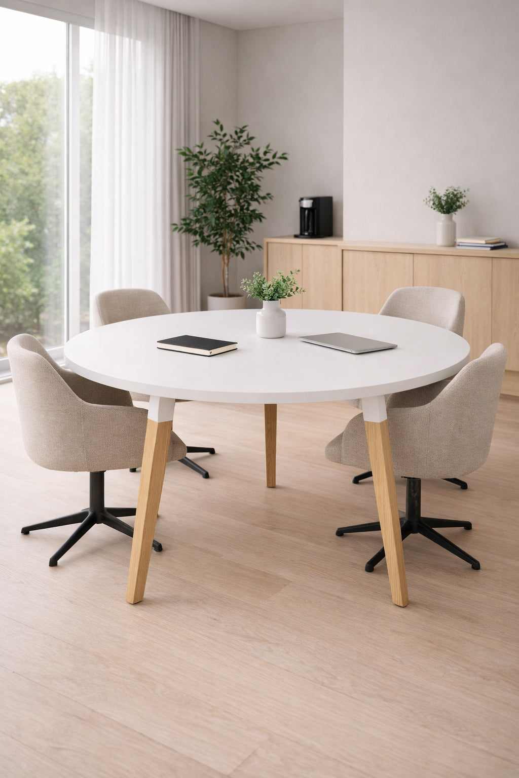 Une salle de réunion moderne avec la Table Réunion Ronde Oslo de Monsieur Bureau, quatre chaises pivotantes beiges, une plante en pot, des cahiers et une fenêtre lumineuse. Une plante et une cafetière sur un buffet en bois clair - idéal pour les espaces de coworking.