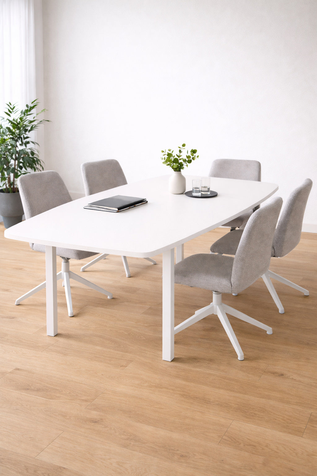 Une salle à manger moderne présente Monsieur Bureau Table de réunion tonneau professionnel Astral 6 pers, six chaises rembourrées grises, un ordinateur portable, un bloc-notes, deux verres et de petites plantes en pot. Un parquet en bois clair et une plante verte en coin complètent le décor.#combo_blanc_blanc