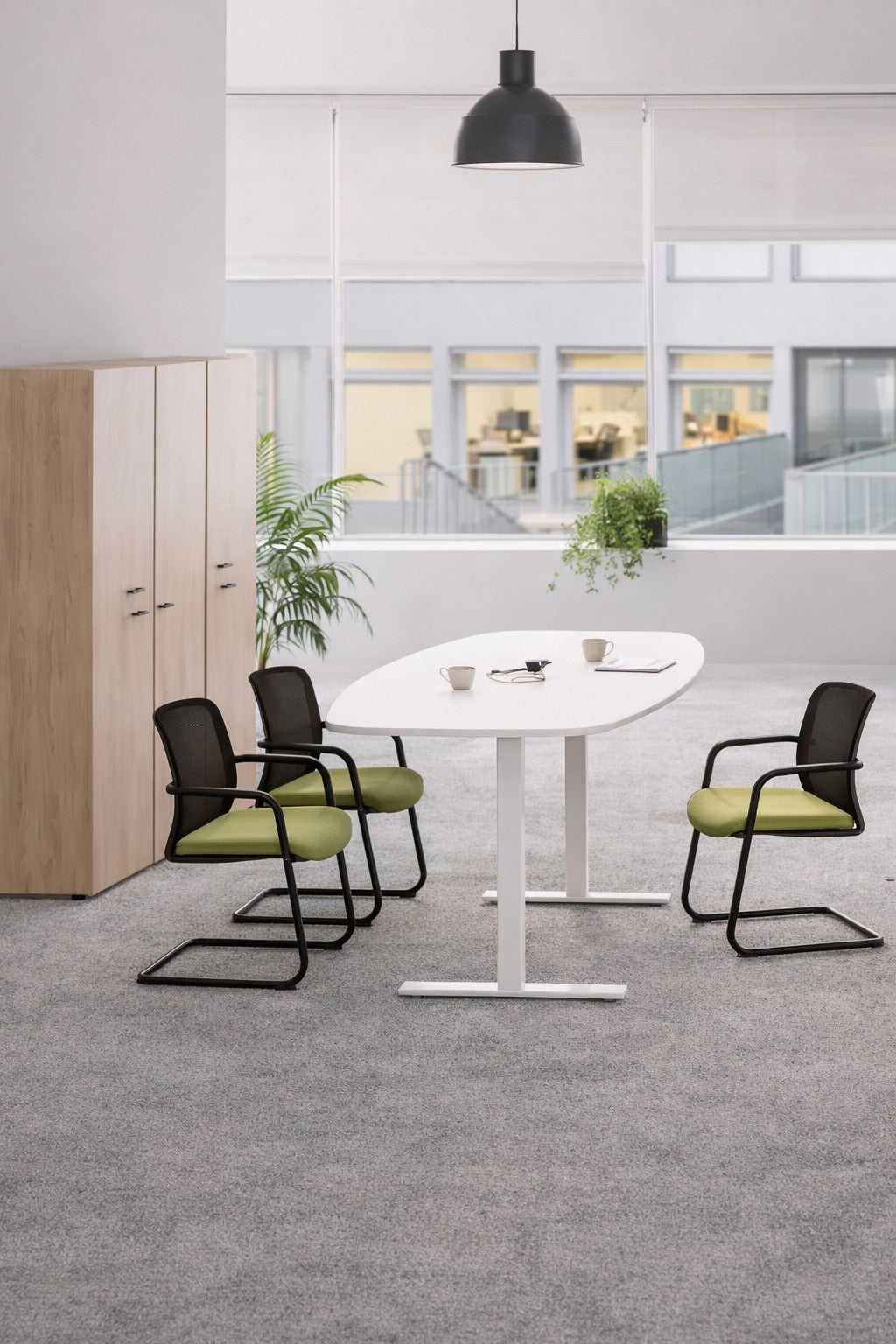 Un espace de réunion moderne meublé avec la Table tonneau électrique Top & Down de Monsieur Bureau pour 6/8 personnes, trois chaises noires avec des sièges verts, un meuble en bois, des plantes en pot et de grandes fenêtres. Deux tasses et un cahier sont posés sur la table.#combo_blanc_blanc
