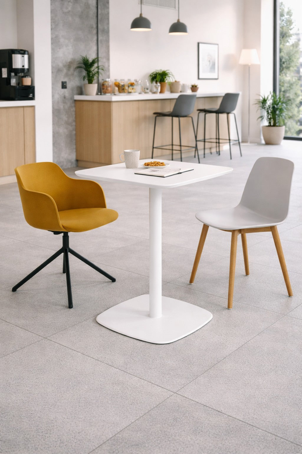 Dans un café moderne, la Table de collectivité Kafeta - Plateau MDF de Monsieur Bureau est associée à une chaise pivotante jaune et à une chaise blanche avec des pieds en bois. Sur la table : une tasse, des biscuits et un livre. Un comptoir et des tabourets de bar sont placés à l'arrière-plan pour un repas décontracté.