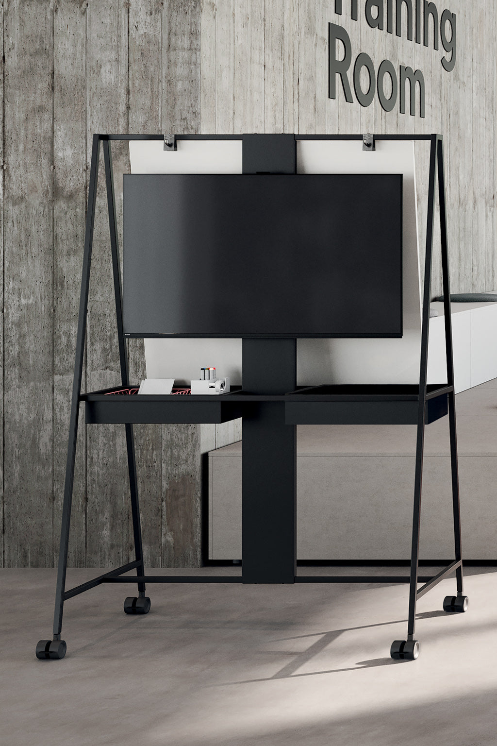 Le Monsieur Bureau Tableau mobile professionnel Agile Hybride, doté d'un grand écran plat, d'un support VESA et d'une étagère pour les marqueurs et les gommes, se dresse sur des pieds à roulettes devant un mur en béton étiqueté Salle de formation.