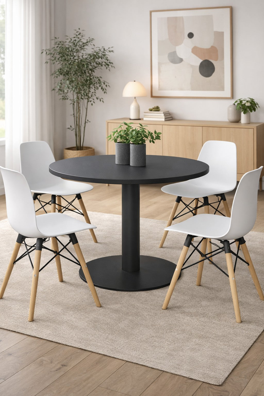 Une salle à manger lumineuse et minimaliste comprenant la Table de Collectivité Ronde Tempo - Plateau Stratifié de Monsieur Bureau, quatre chaises blanches avec des pieds en bois, un tapis beige, un buffet, une plante en pot et de l'art mural abstrait.#combo_graphite_graphite